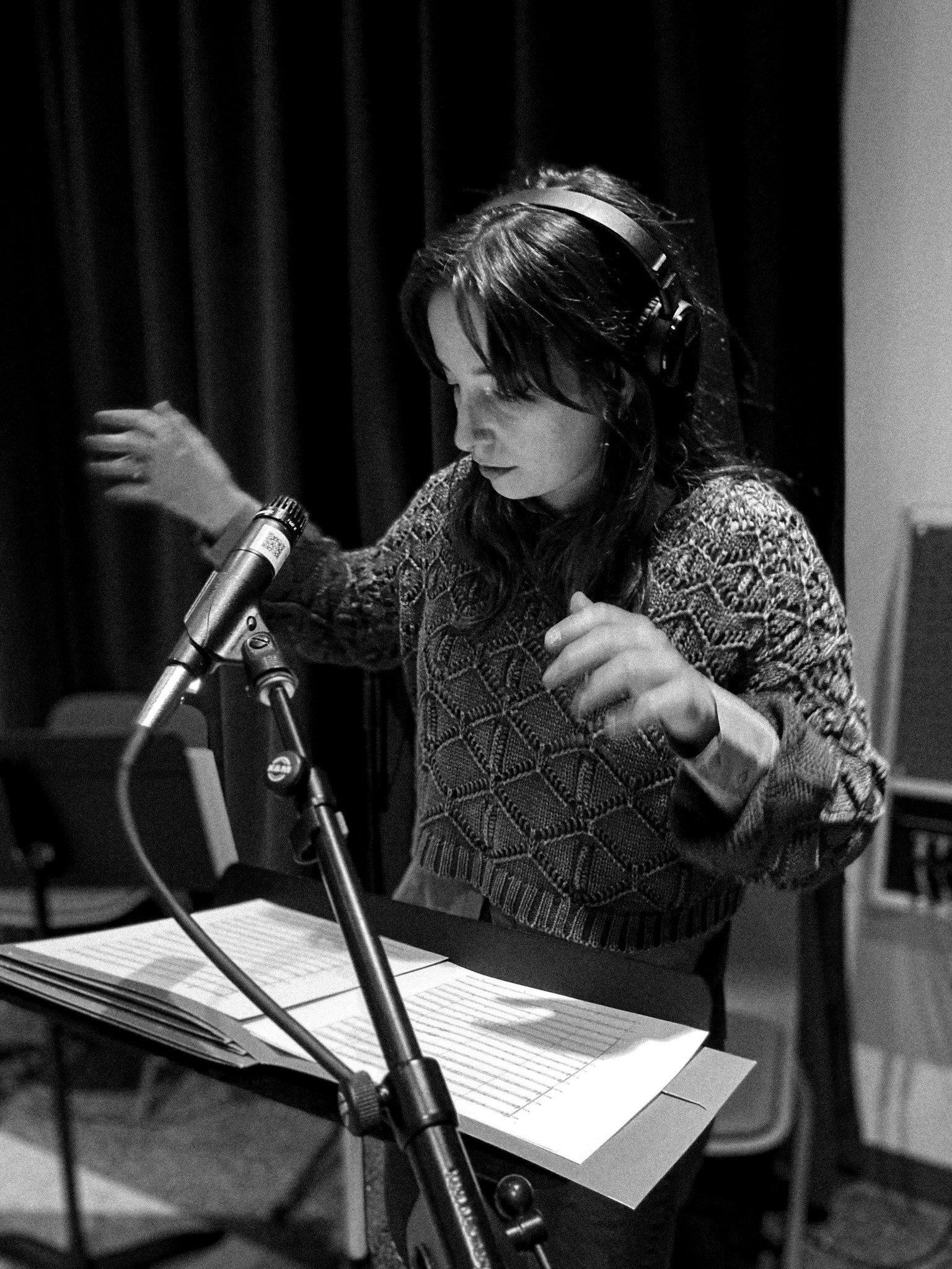 Woman wearing headphones, standing at a music stand with sheet music, speaking into a microphone in a recording studio.