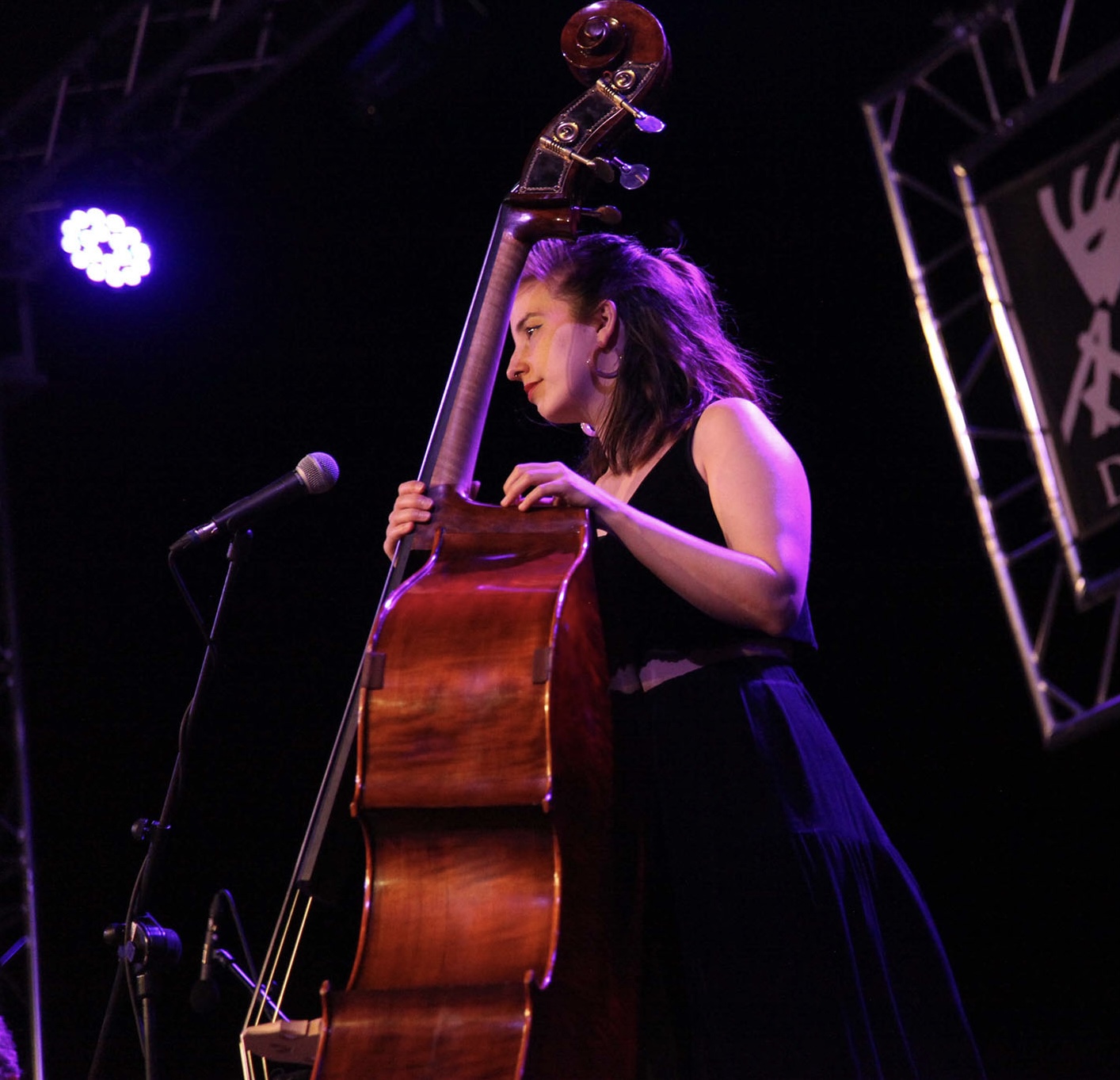 Young woman playing double bass on stage with purple lighting and microphone nearby.