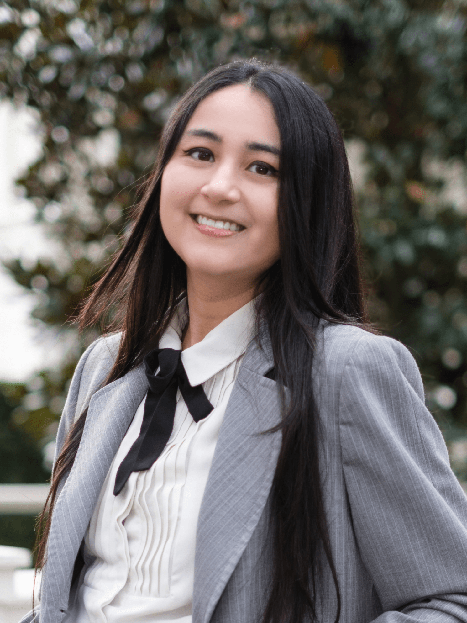 Young woman with long dark hair smiling outdoors, wearing a gray blazer and white shirt with a black tie.