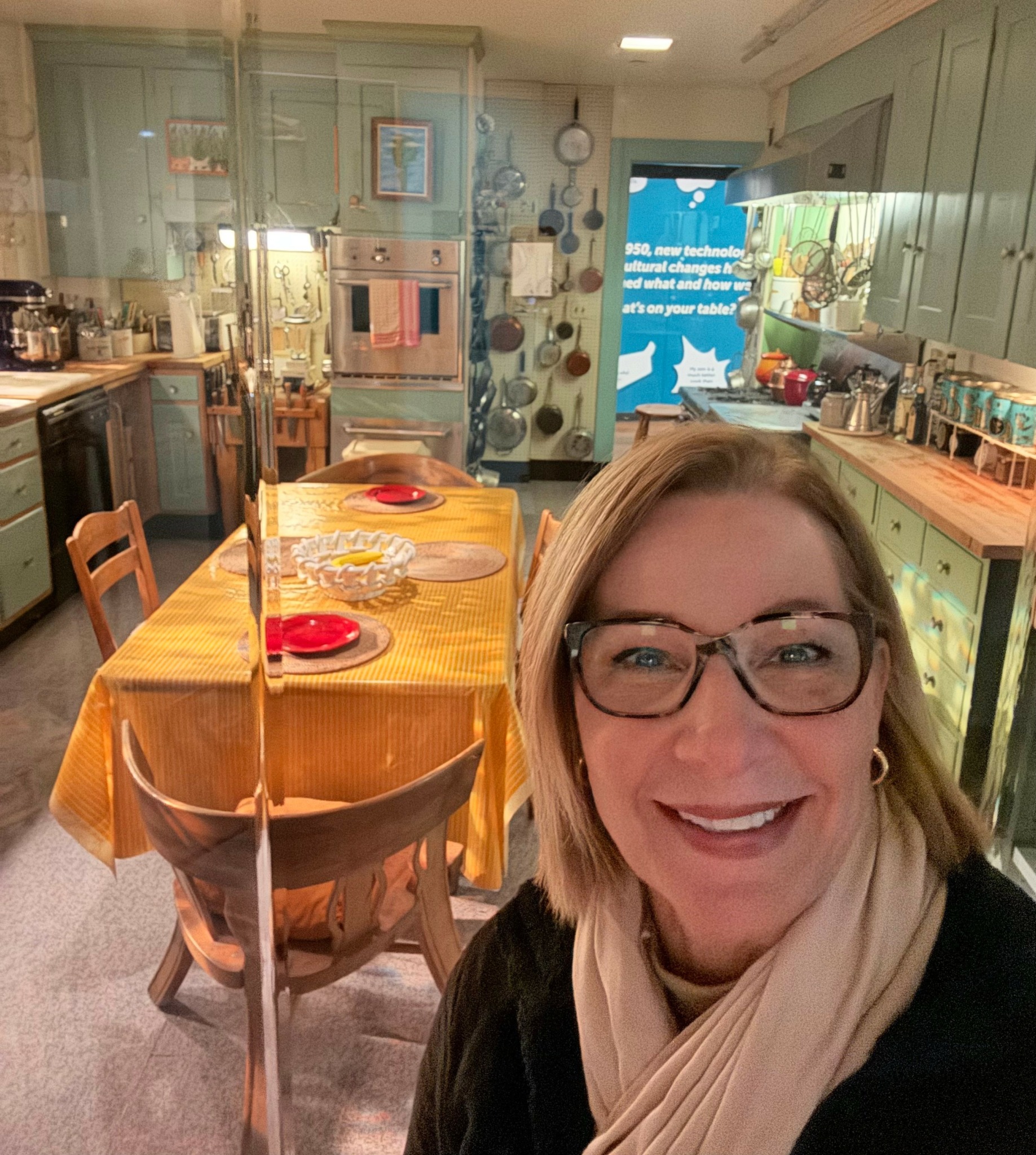 Woman with glasses smiling in a kitchen with a dining table set with red plates and a yellow tablecloth.