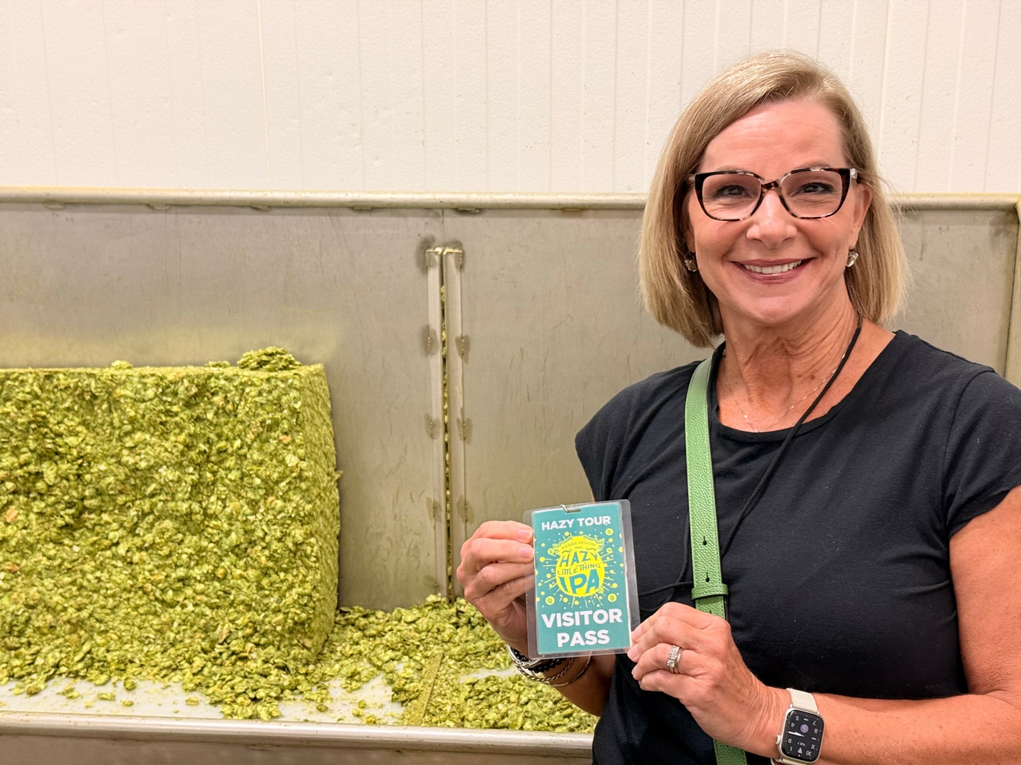 Smiling woman holding a visitor pass, standing next to a large pile of green hops, in an indoor setting.