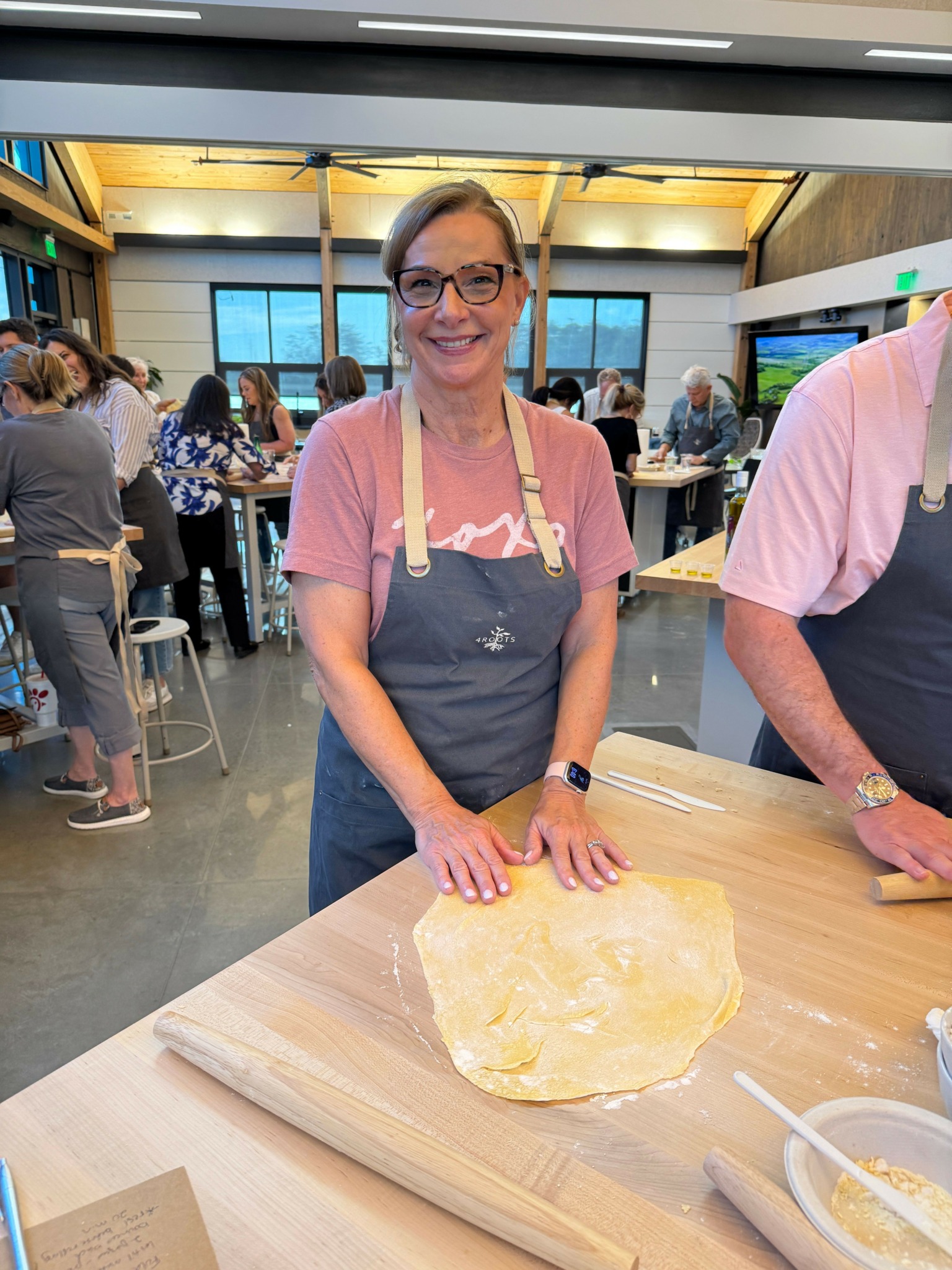 Woman with glasses and a pink shirt rolls dough on a table in a busy kitchen or cooking class setting.