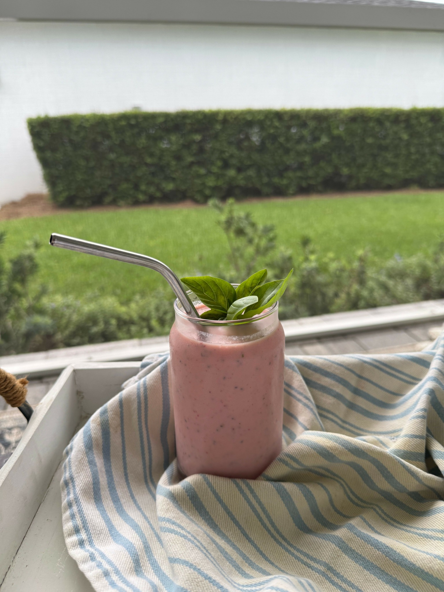 Pink smoothie in a glass with a metal straw, garnished with green leaves, on a striped cloth near a window.