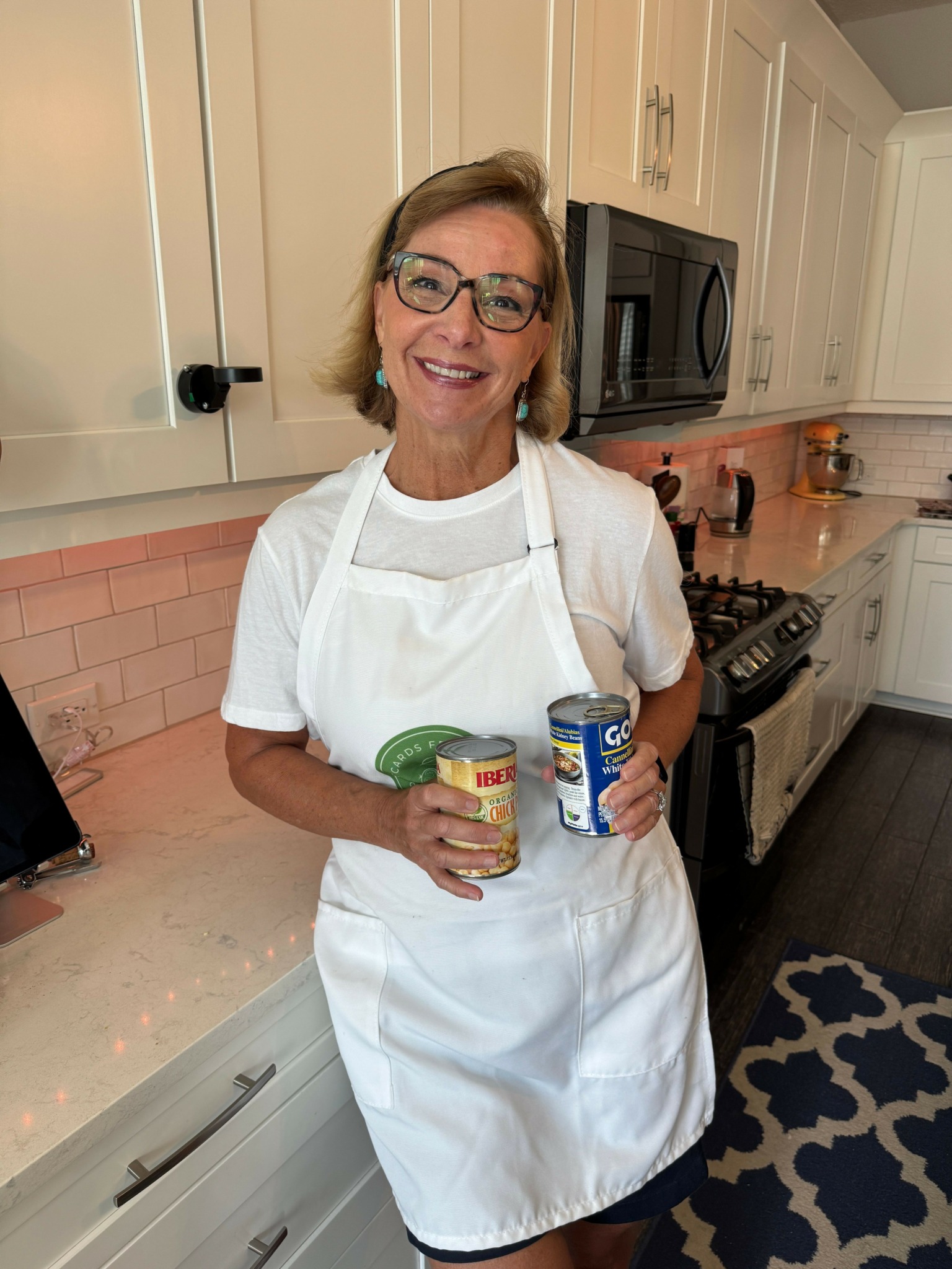 Woman with glasses in a white apron holding a jar and a can in a kitchen.