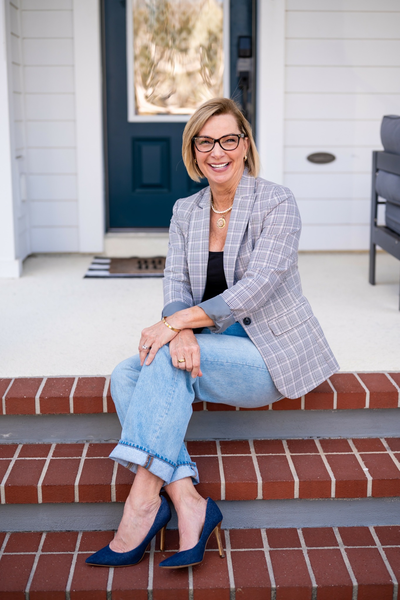 Woman sitting on front porch steps, smiling, wearing glasses, blazer, jeans, and high heels.