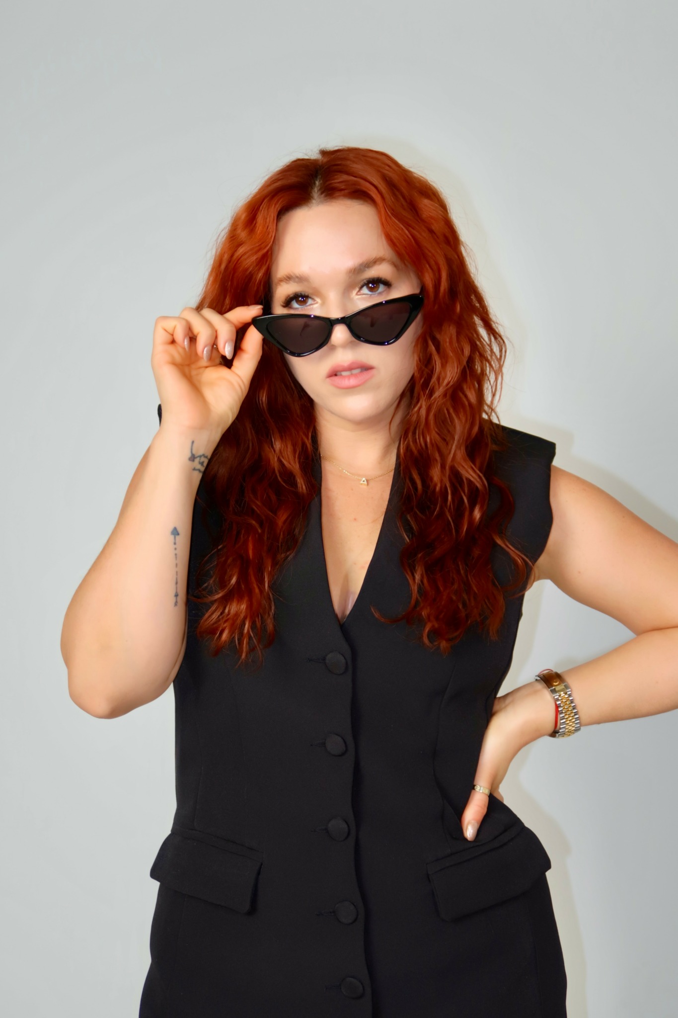 Woman with red hair wearing sunglasses and a black sleeveless vest, posing with one hand on her hip.