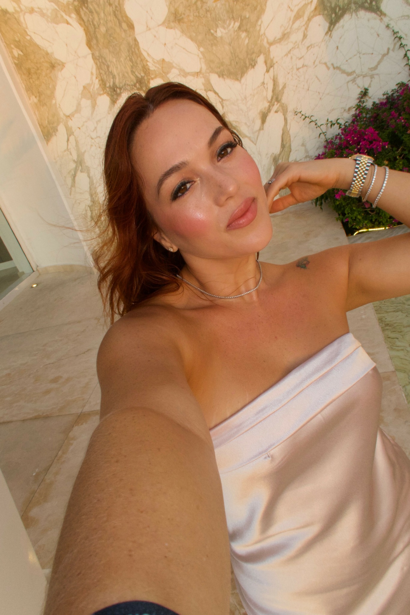 Woman with shoulder-length hair taking a selfie outdoors, wearing a strapless white top, with a stone wall and flowers in background.