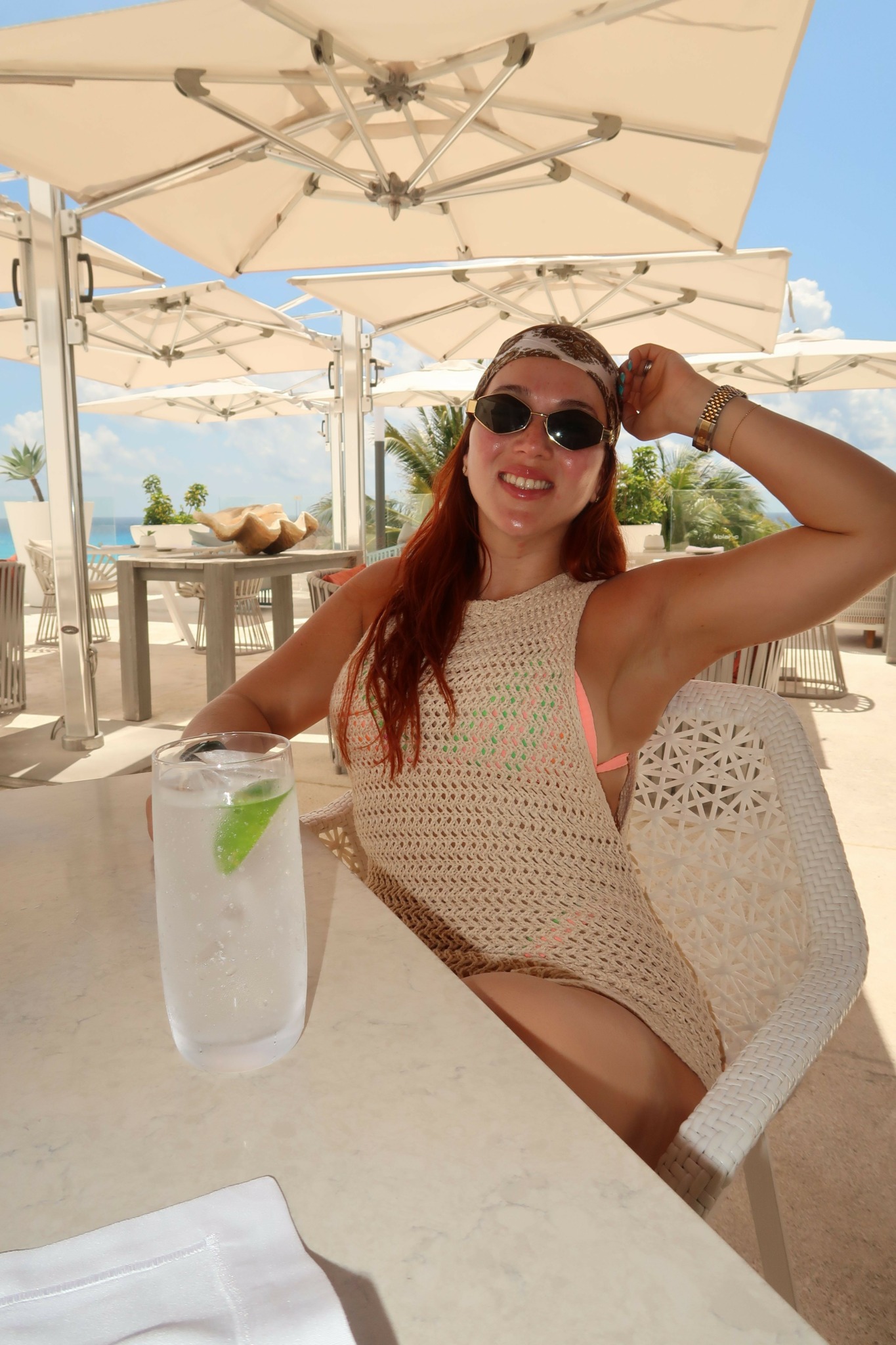 Woman in sunglasses and a beige dress sitting at an outdoor table with a drink, under a large umbrella.