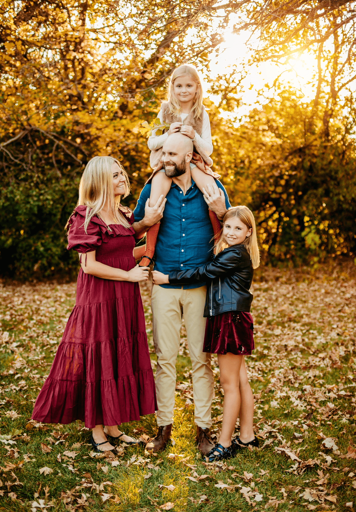 Four people outdoors in autumn, one child sitting on a man's shoulders, three standing around him, trees and sunlight in background.