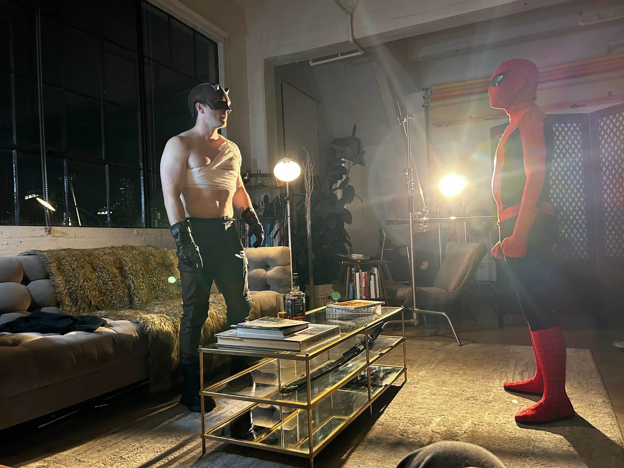 Person dressed as Spider-Man facing another person in a living room with a sofa, coffee table, and large windows.