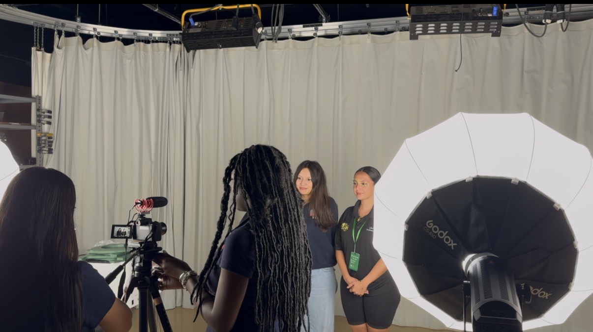 Two women stand in front of a white backdrop with a large studio light and camera, preparing for a photo or video shoot.