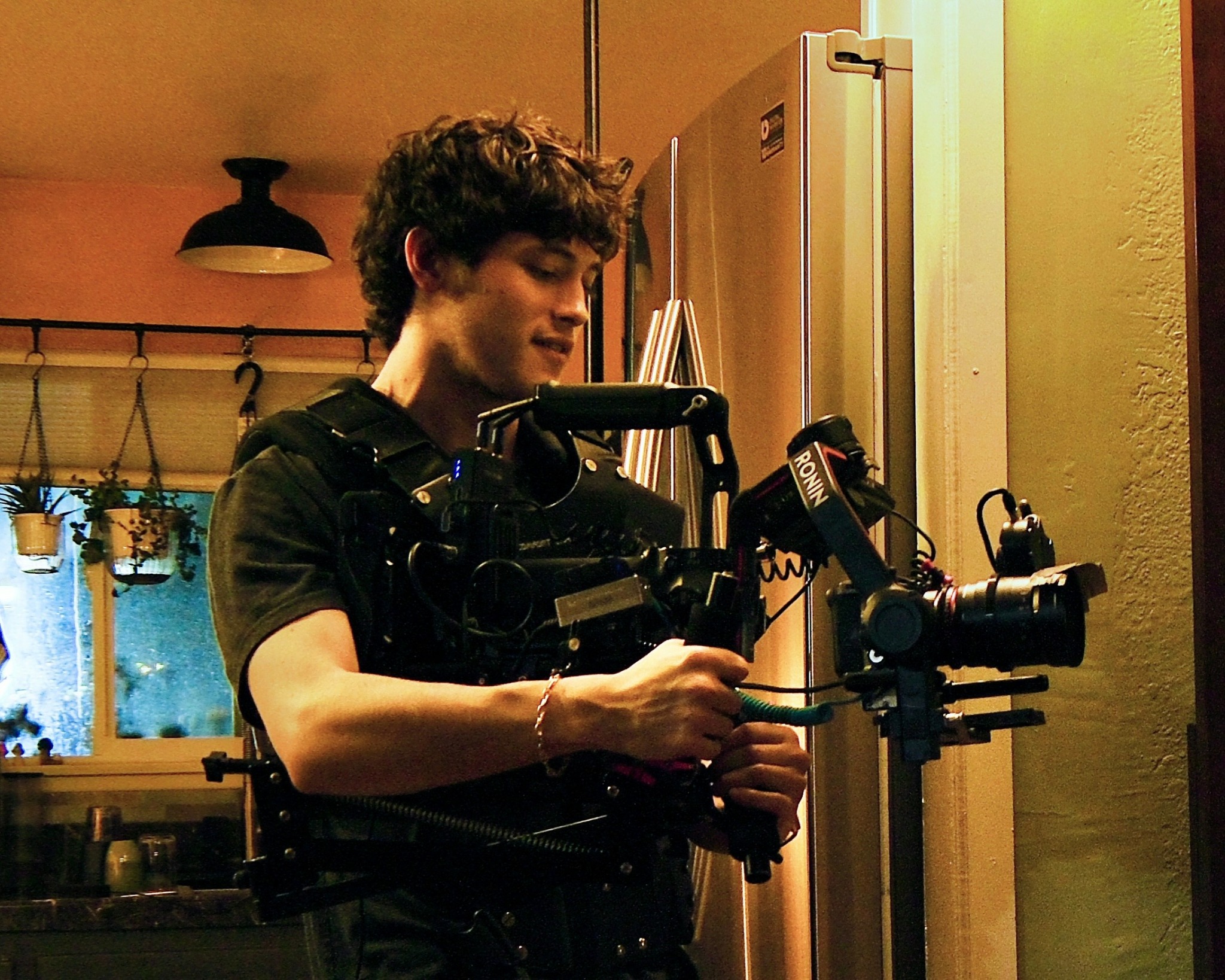 Young person operating a professional camera rig indoors, with kitchen background and warm lighting.