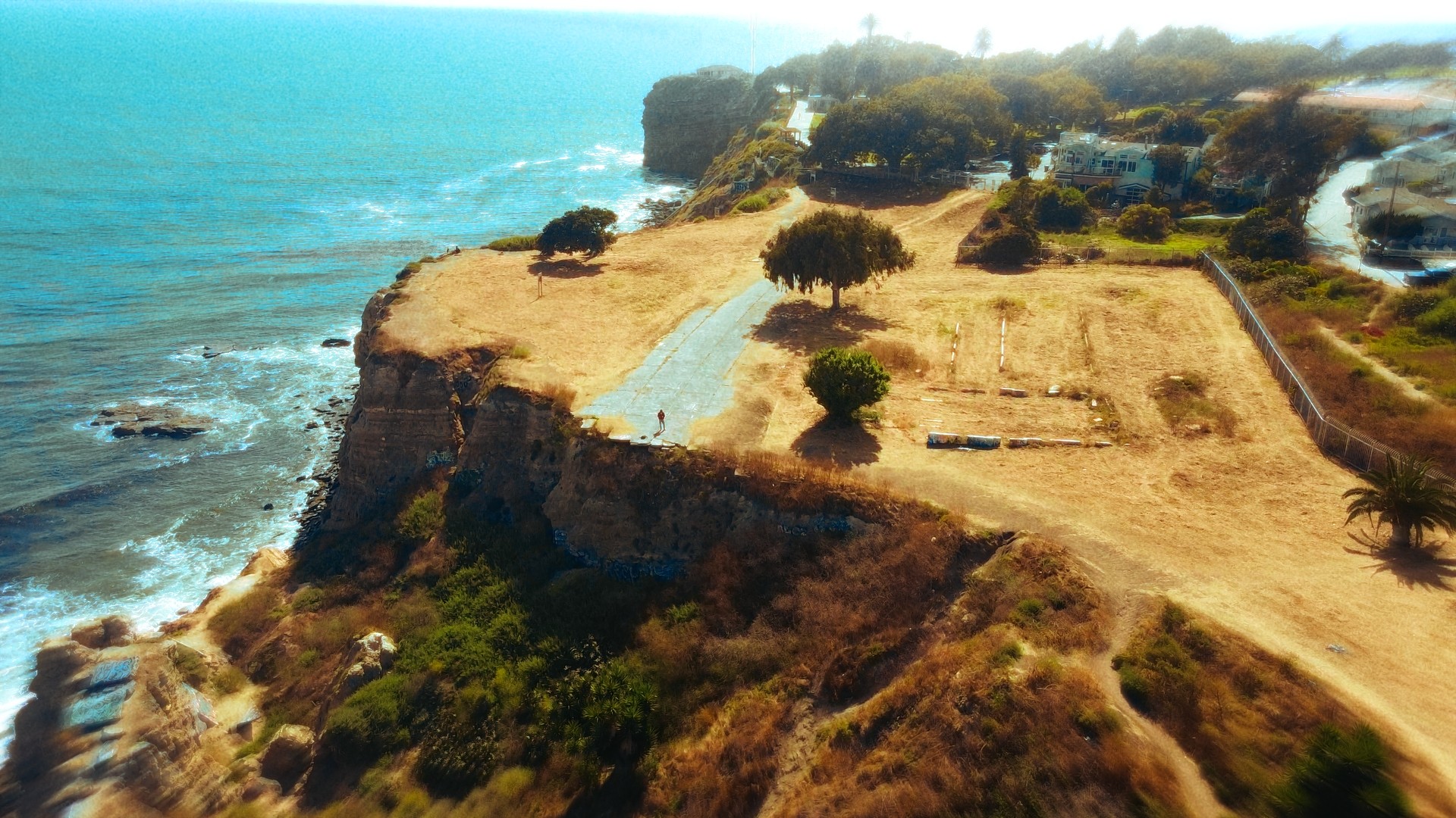 Cliffside overlooking ocean with trees and grassy areas, coastline extending into distance, bright sky, and waves crashing below.