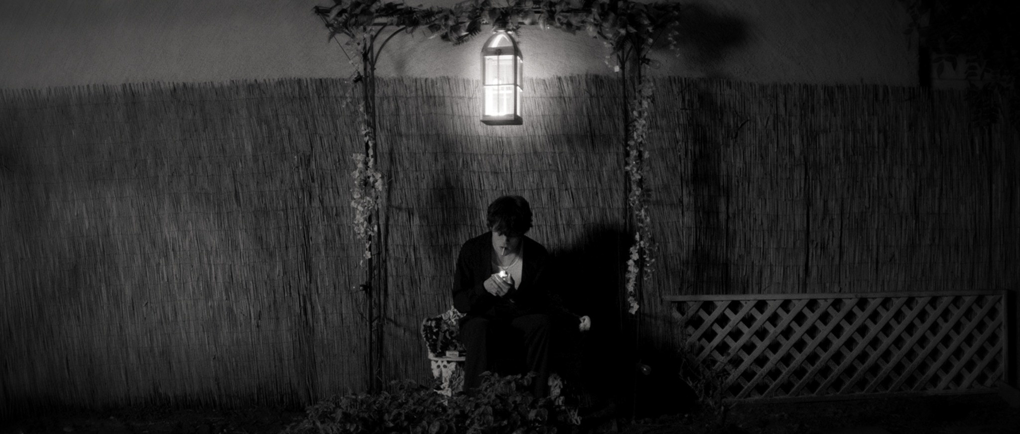 Person sitting on ground under a hanging lantern at night, with a wooden fence and lattice in background.