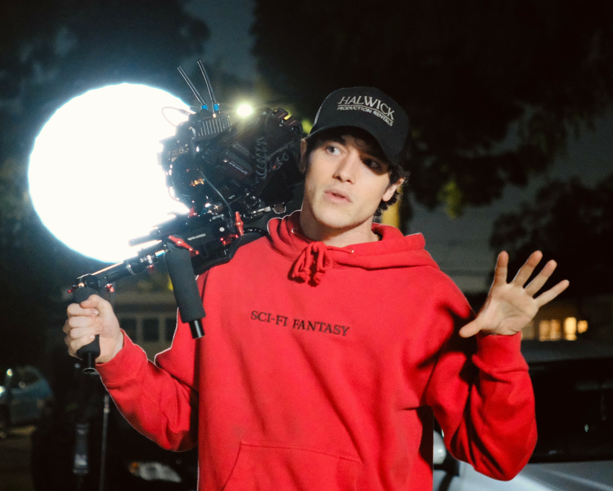 Person in red hoodie and black cap holding camera with bright light behind outdoors at night.