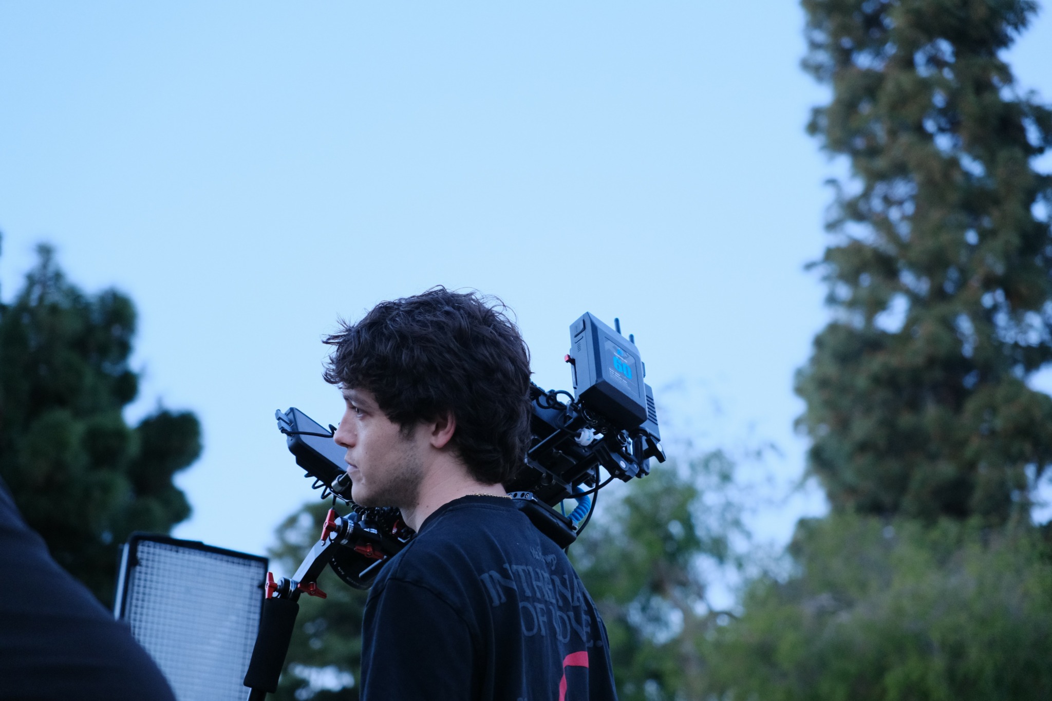 Young man with curly hair outdoors, carrying camera equipment on his shoulder, trees in background.