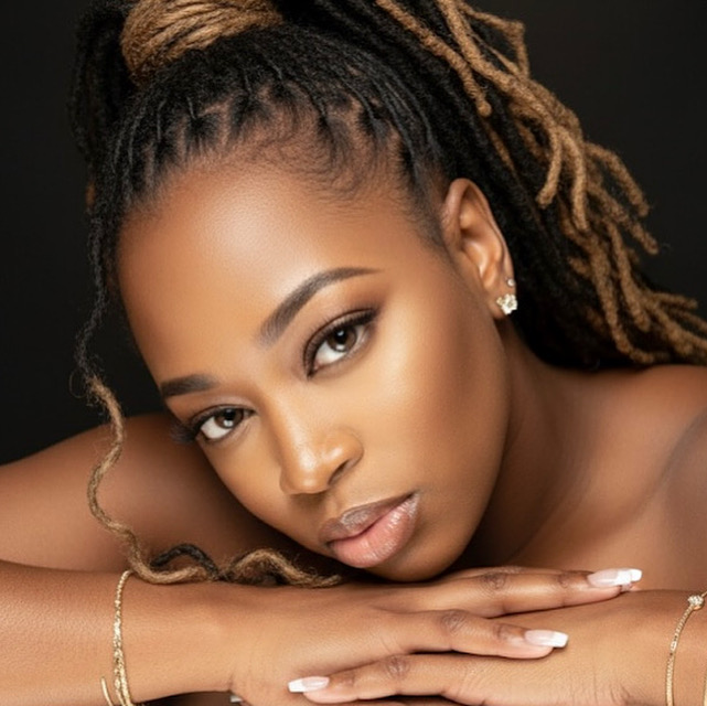 Close-up of a woman with styled dreadlocks, resting her head on her arms, looking at the camera.