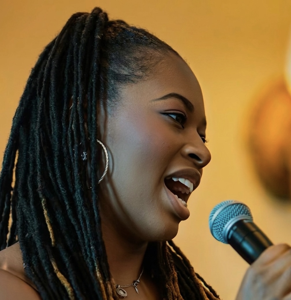Woman with dreadlocks singing into a microphone, smiling, against a yellow background.