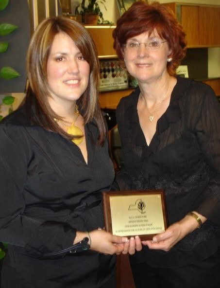 Two women standing indoors, holding a framed certificate or award, smiling at the camera.