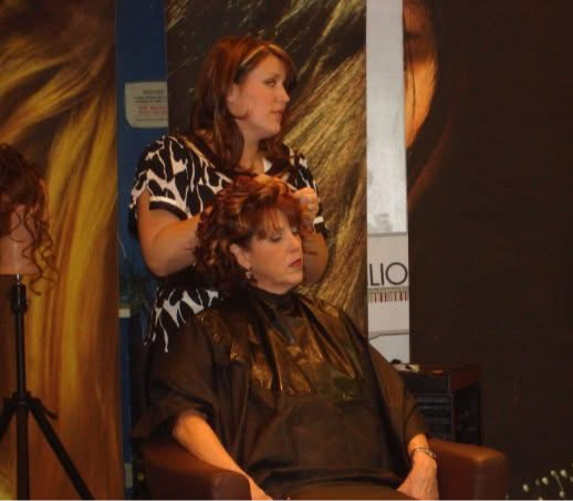 Woman getting her hair styled by a hairdresser in a salon, with a poster in the background.
