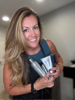 Woman with long blonde hair smiling, holding skincare products, in an indoor setting.
