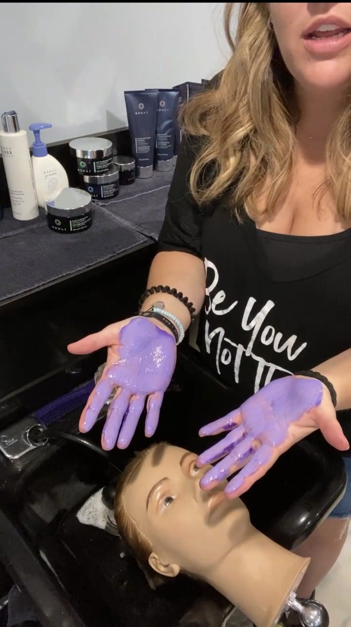 Person wearing purple gloves holds mannequin head with hair, in a salon setting with hair products on counter.