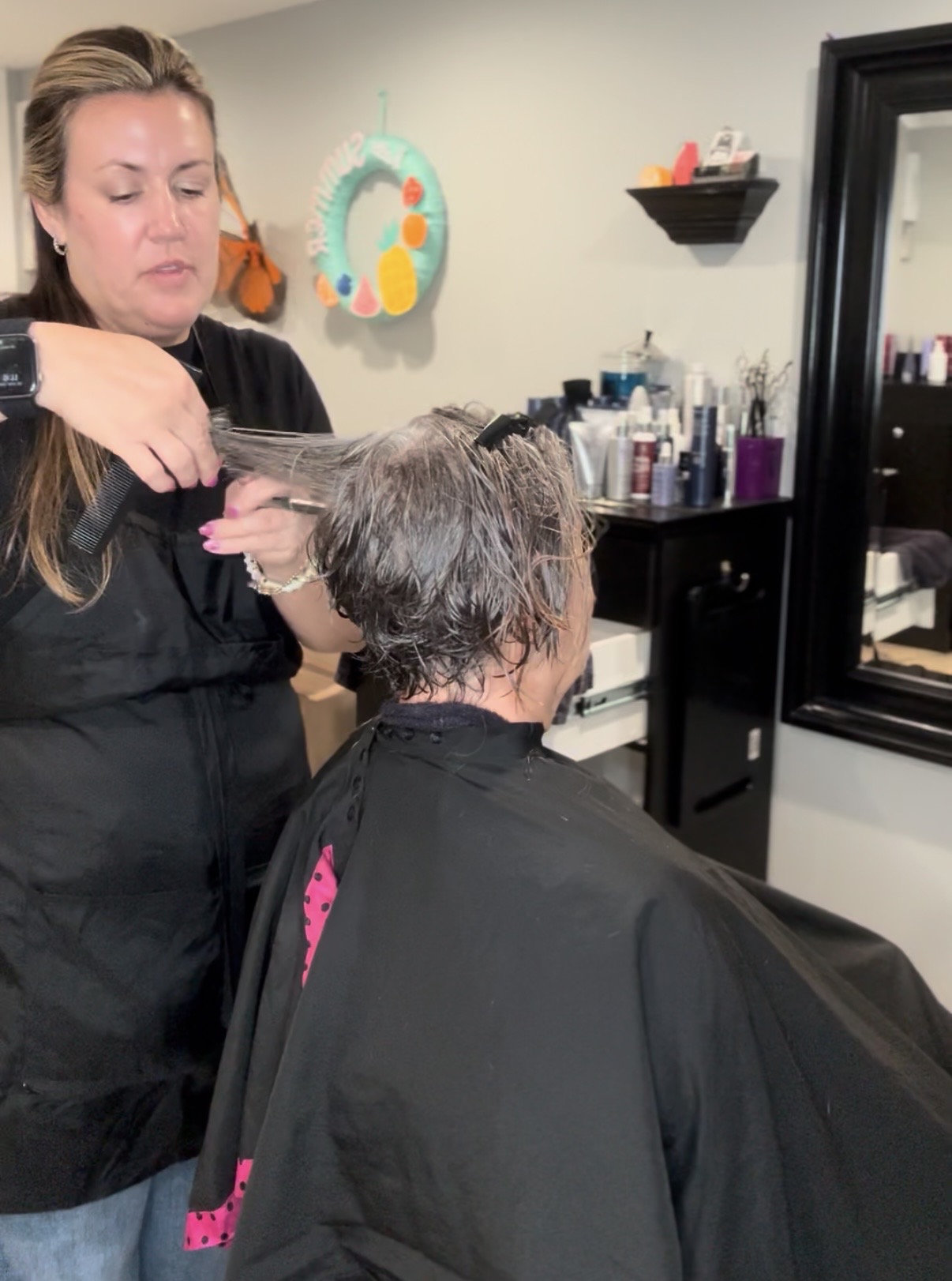 Hairdresser cutting a person's hair in a salon with shelves of products and a mirror in the background.