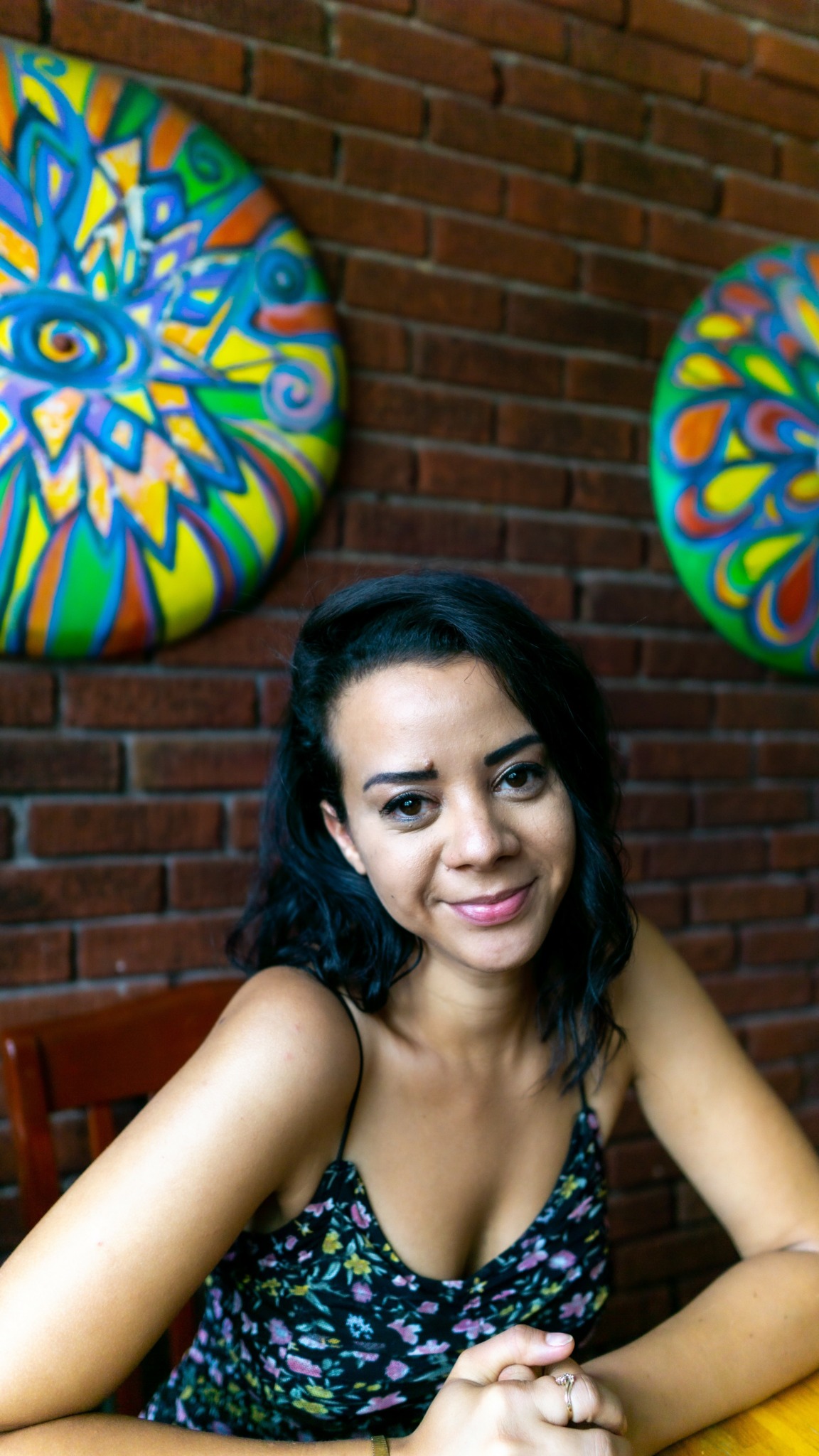 Young woman with dark hair smiling, sitting at a table in front of colorful circular artwork on brick wall.