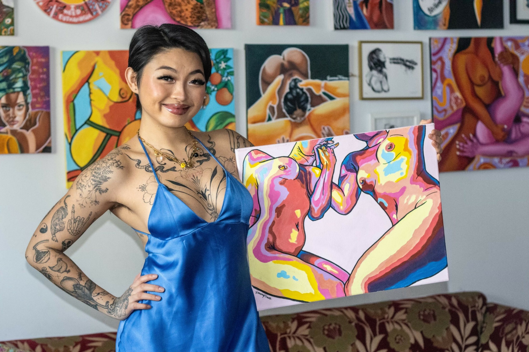 Young woman with tattoos in a blue dress standing in art gallery with colorful paintings and holding a painting.