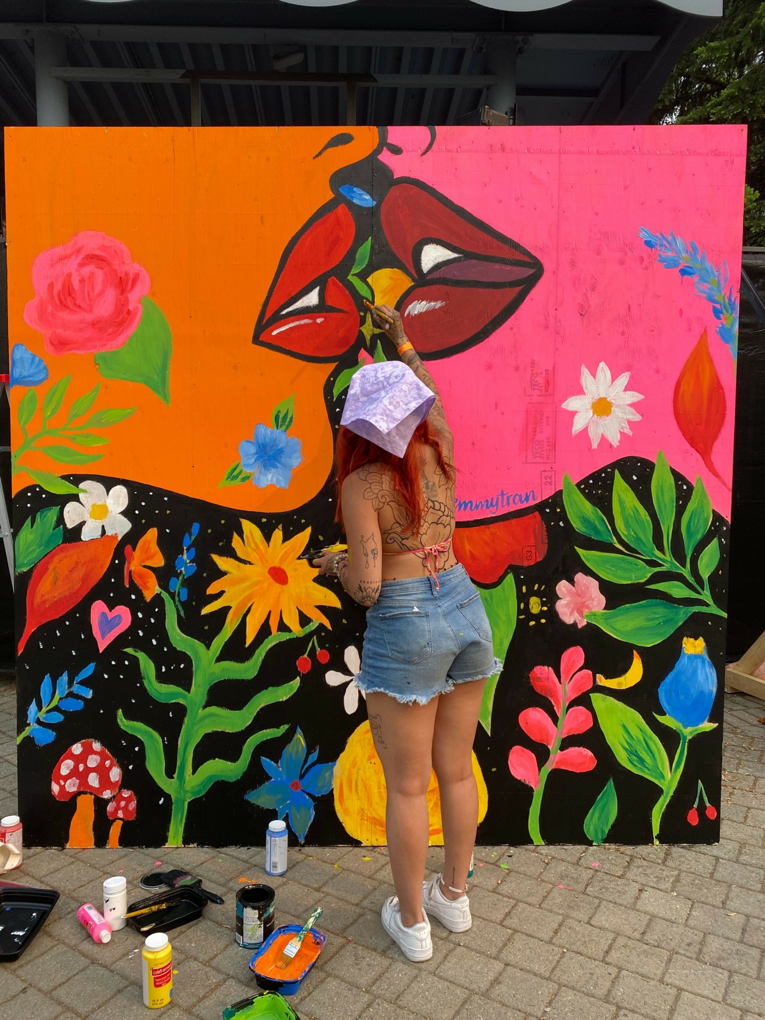 Person lying on colorful painted mural with flowers and abstract shapes, outdoors on paved surface, surrounded by paint supplies.