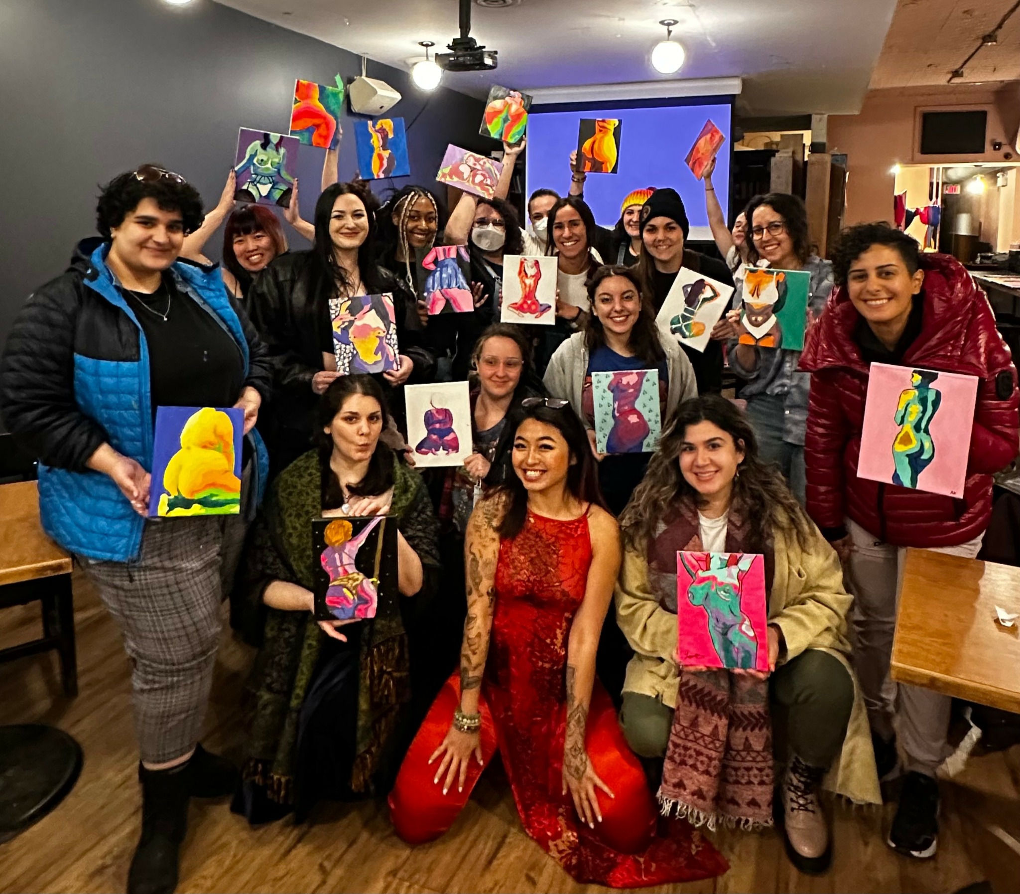Group of people holding colorful paintings in an indoor setting with a large screen in the background.