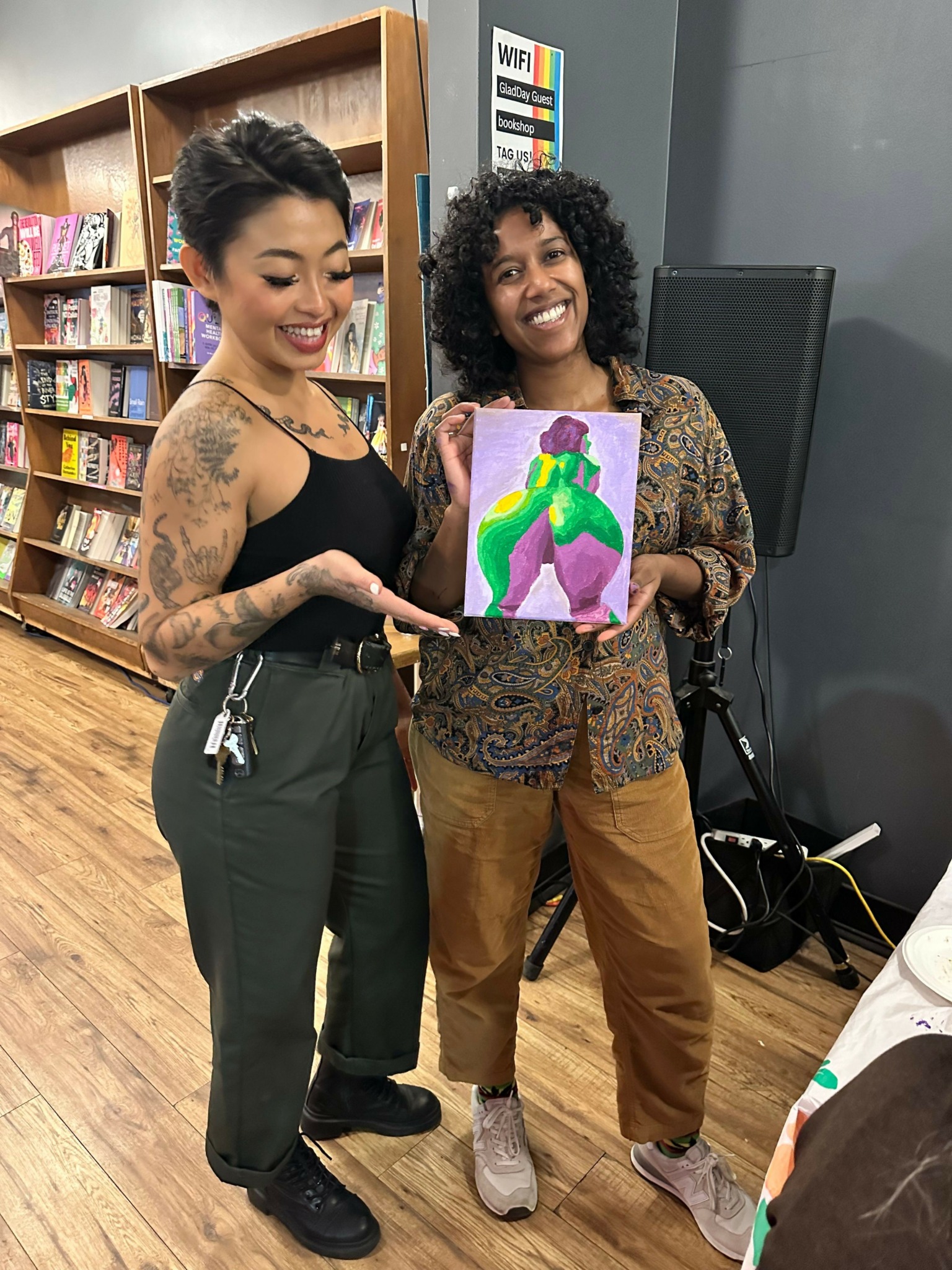 Two women standing in a room, one holding a colorful artwork, shelves with books in the background.