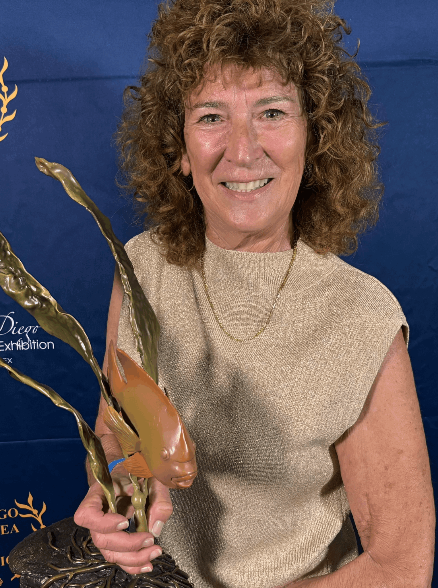 Woman with curly hair holding an award trophy, smiling, against a blue background.