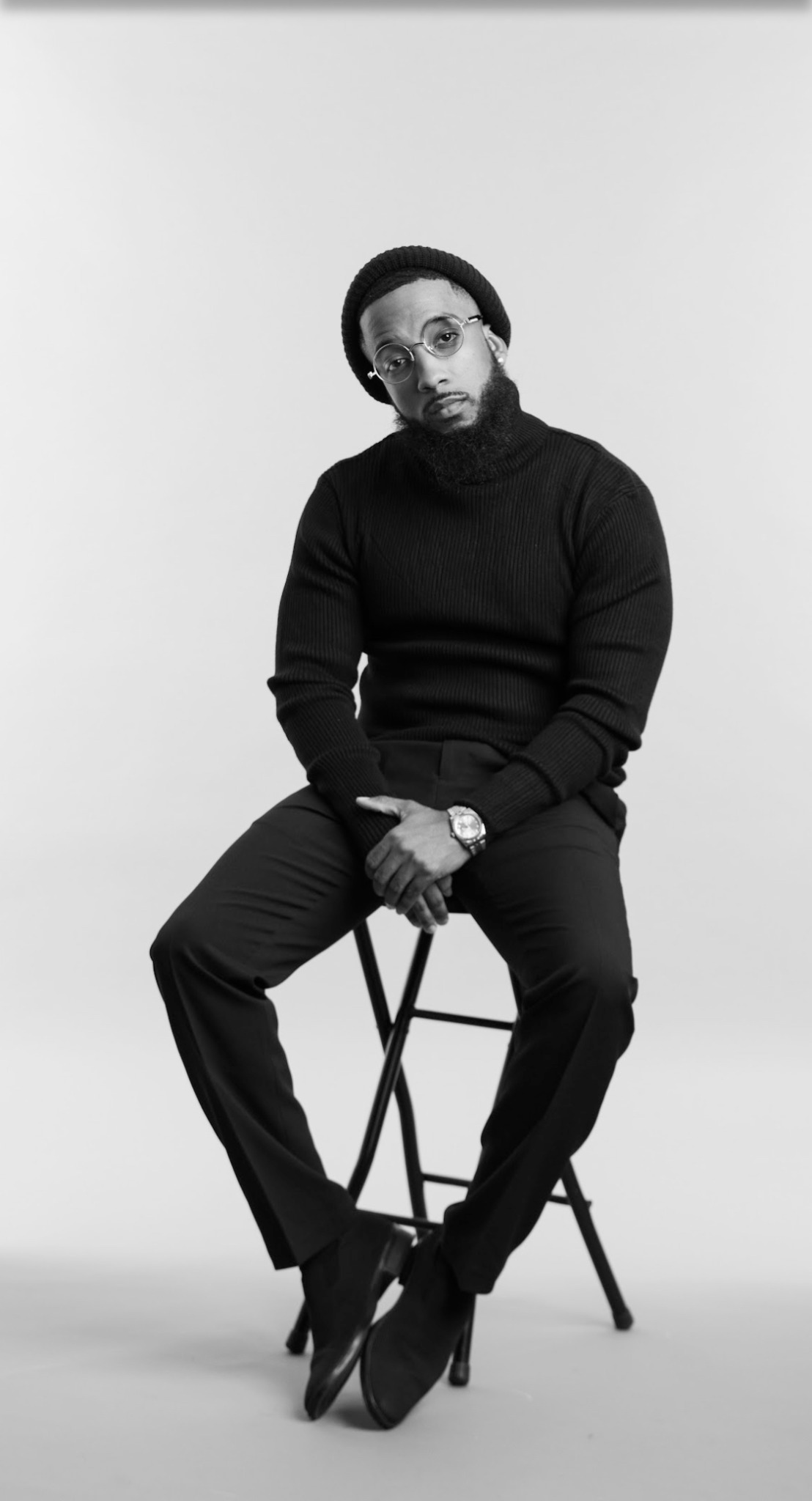 Man sitting on a chair, wearing a black turtleneck, glasses, and a watch, against a plain background.