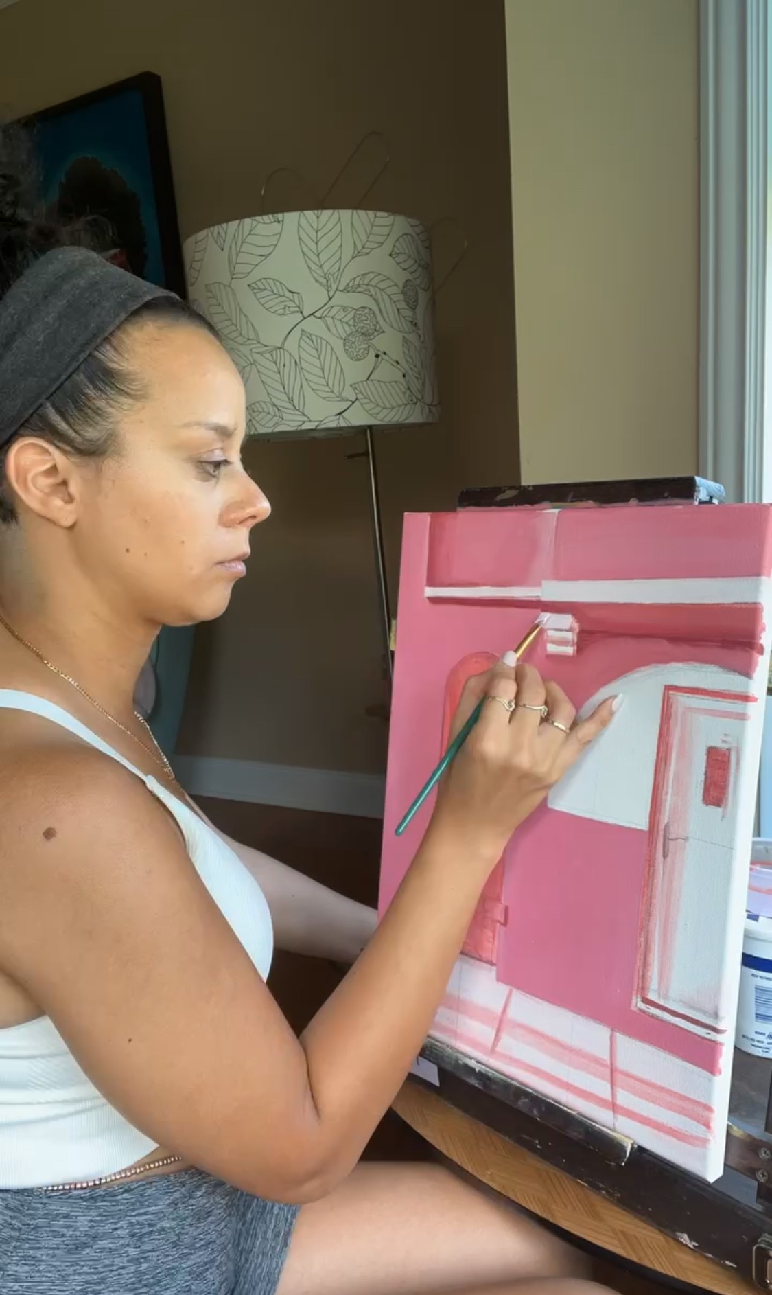 Woman painting on a pink canvas at a table indoors, with a lamp and window nearby.
