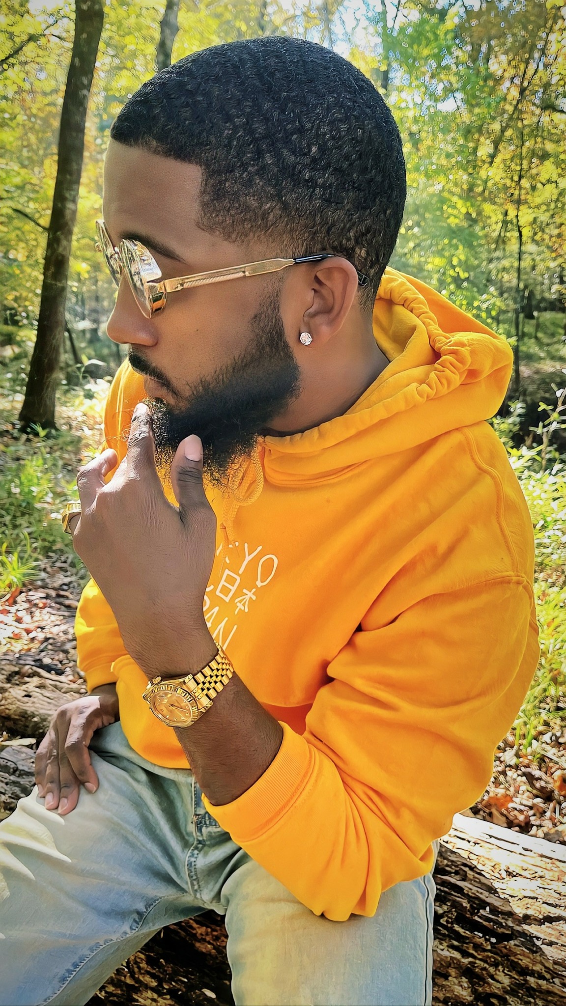 Young man with glasses and a beard, wearing a yellow hoodie, sitting outdoors in a wooded area.