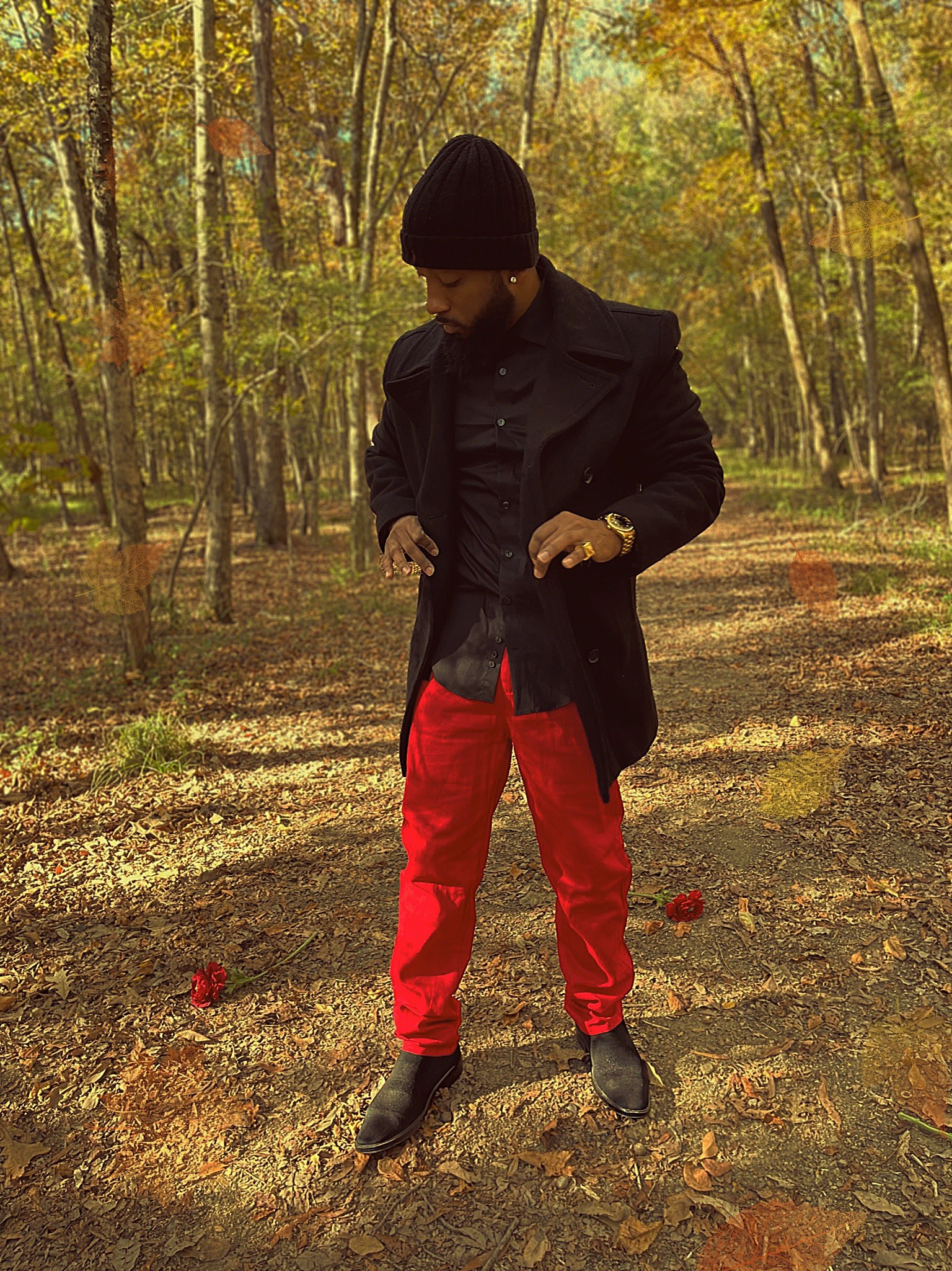 Person standing outdoors in a forest with trees and fallen leaves, wearing a black coat, black hat, and red pants.