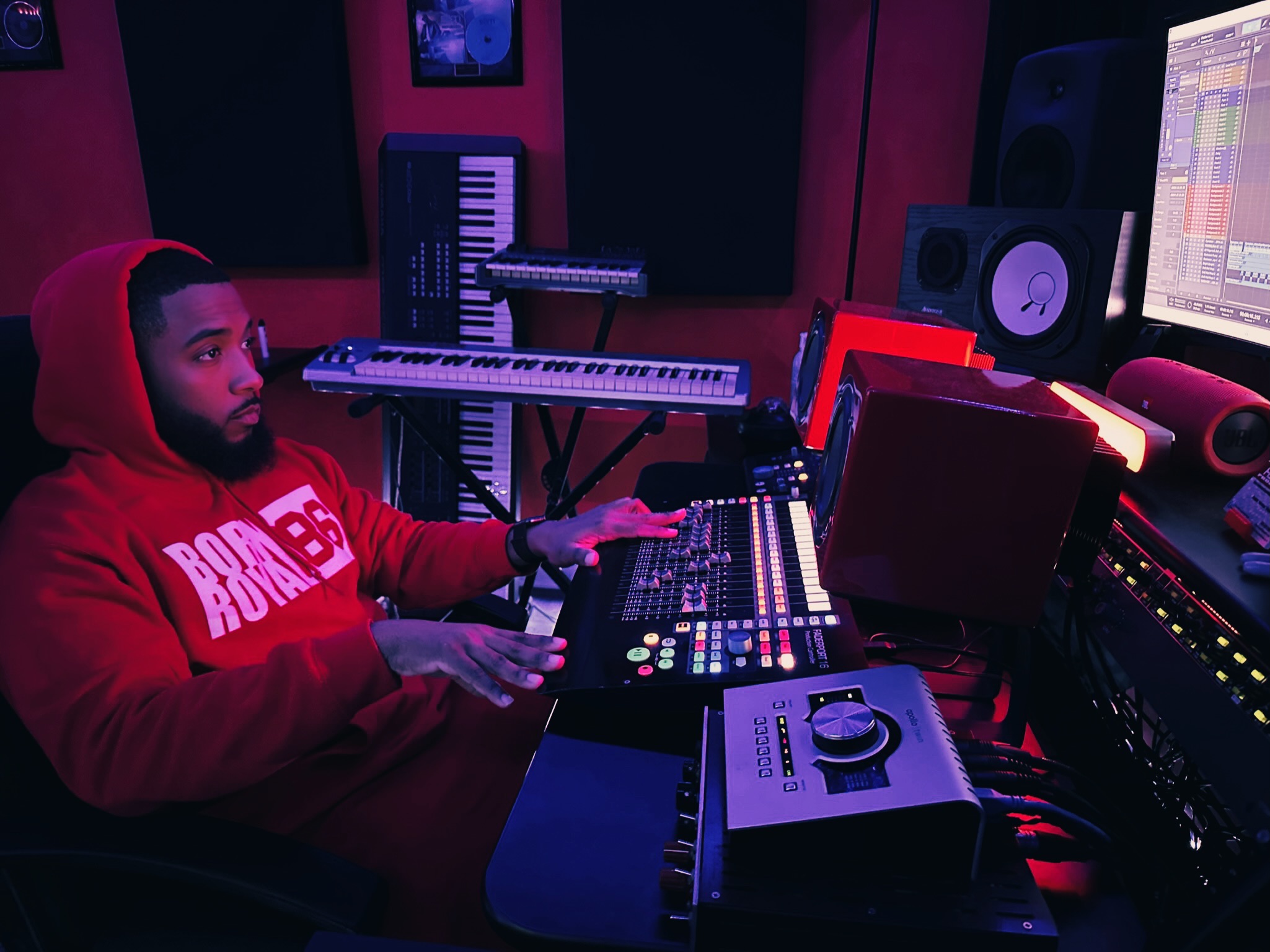 Person in red hoodie working at music production equipment in a studio with electronic instruments and speakers.
