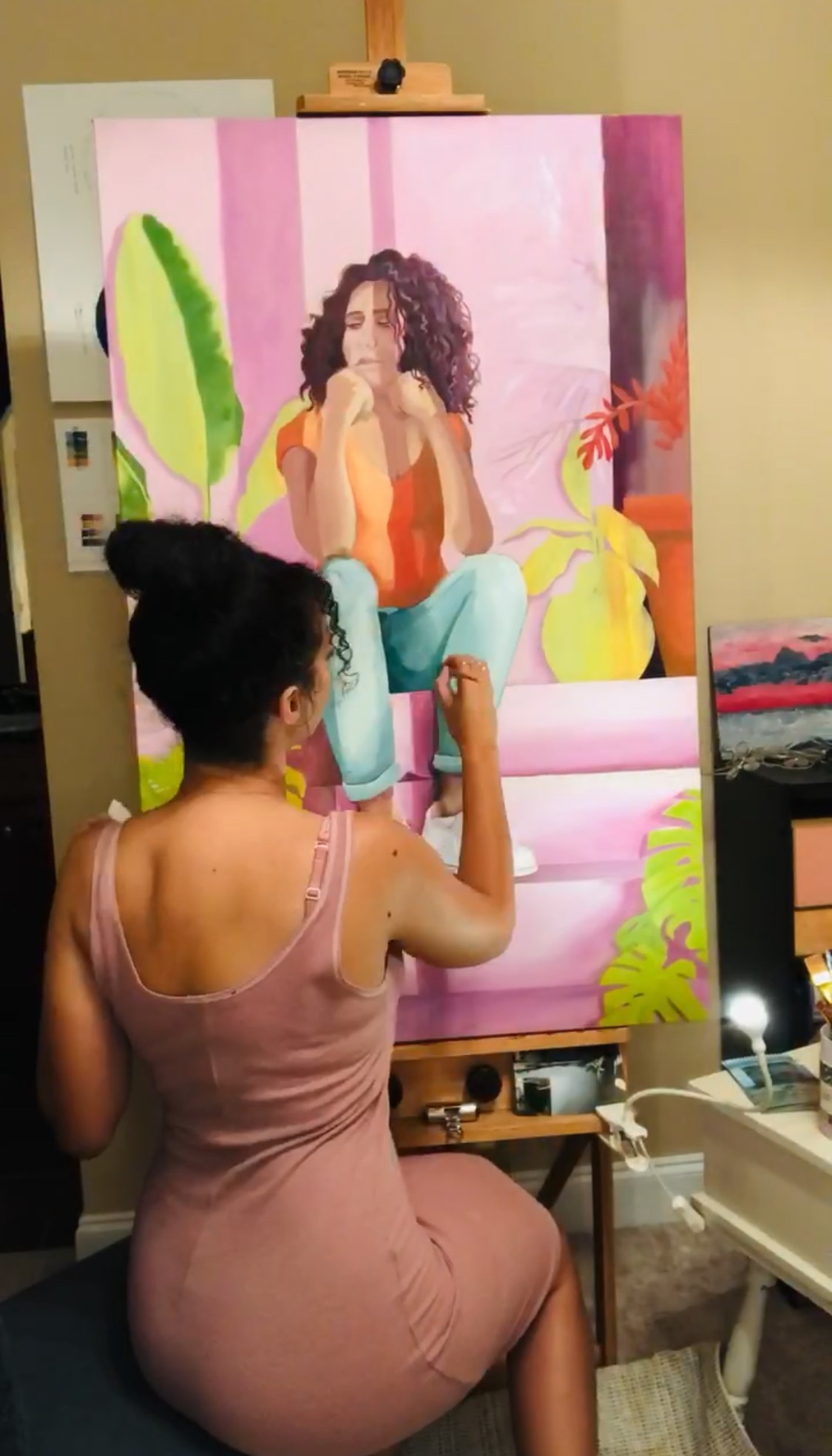 Woman in pink dress painting a colorful portrait of a seated woman with curly hair, plants, and a pink background.