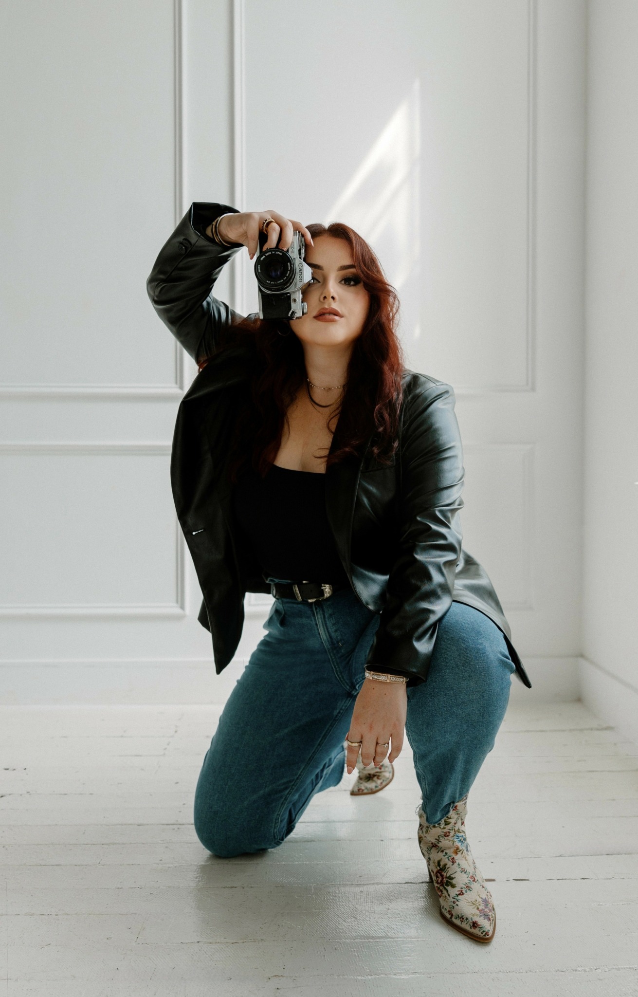 Woman with red hair wearing black leather jacket and blue jeans, holding camera, kneeling indoors.