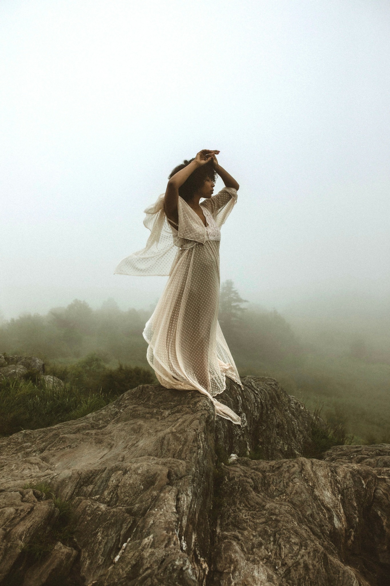 Woman in a flowing dress standing on a rock, with foggy landscape in the background.