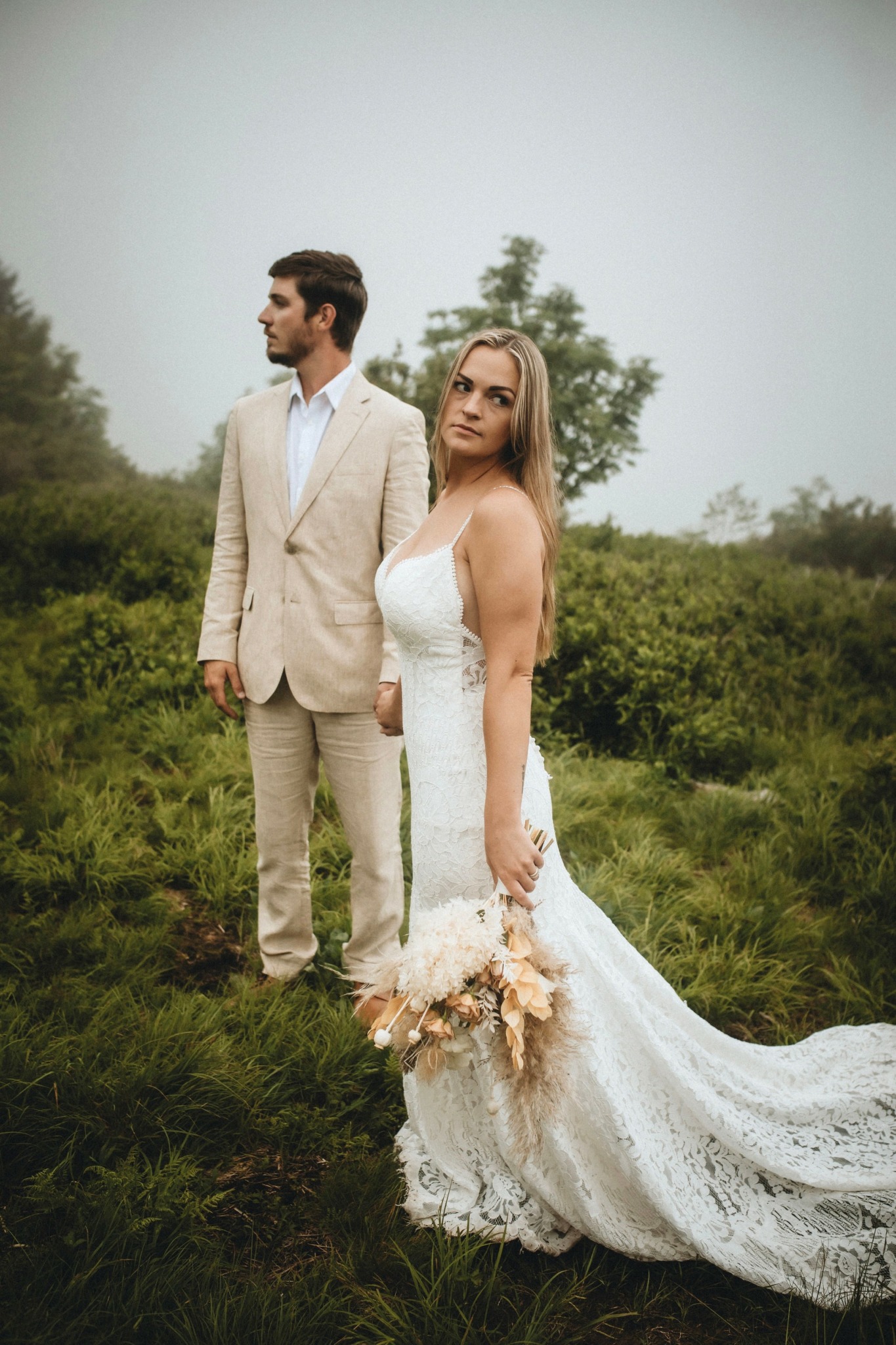Bride in a white lace wedding dress holding a bouquet, standing outdoors with groom in a beige suit behind her.