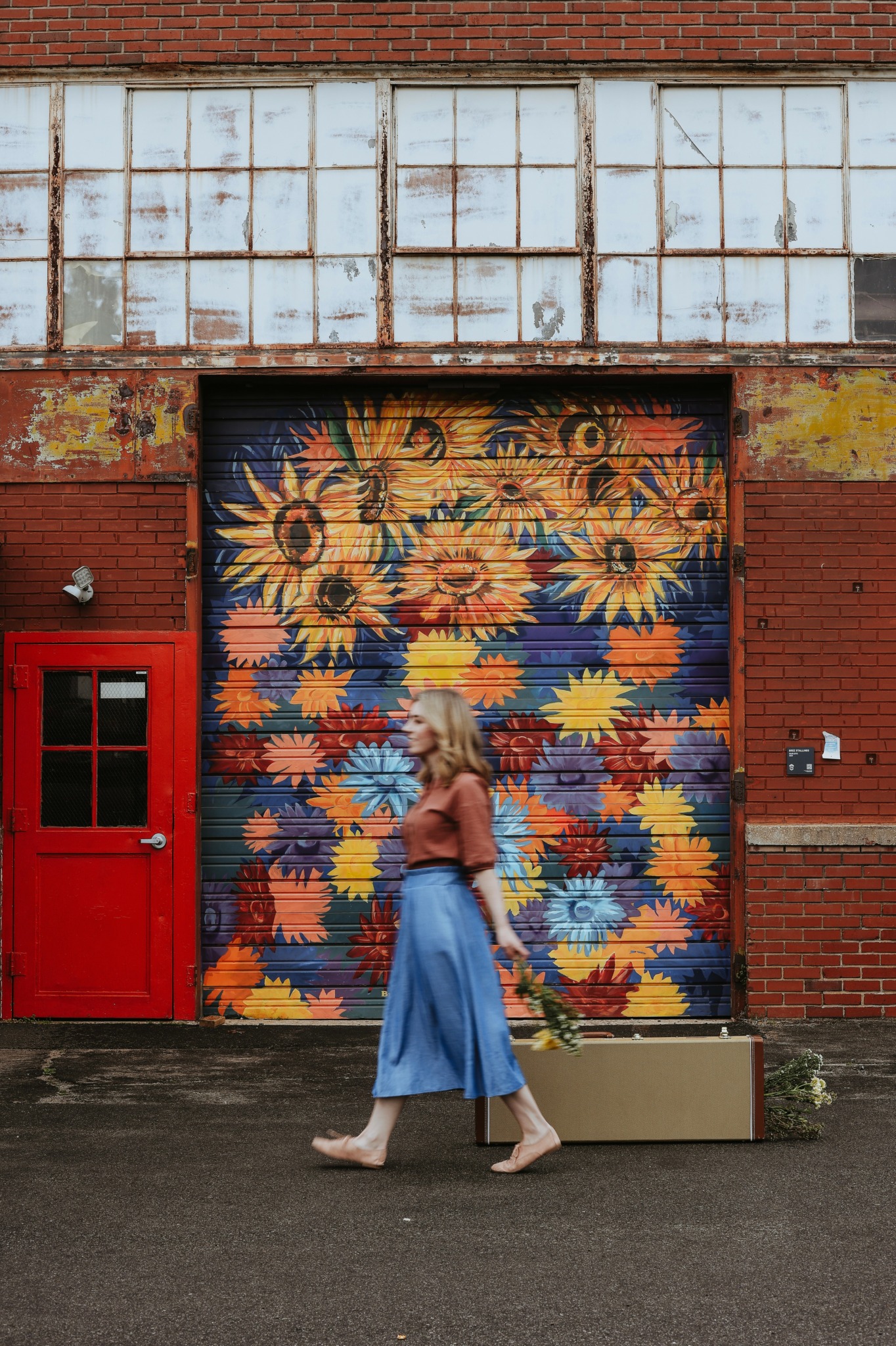Woman walking past a colorful floral mural on a garage door, holding a flower in her hand.