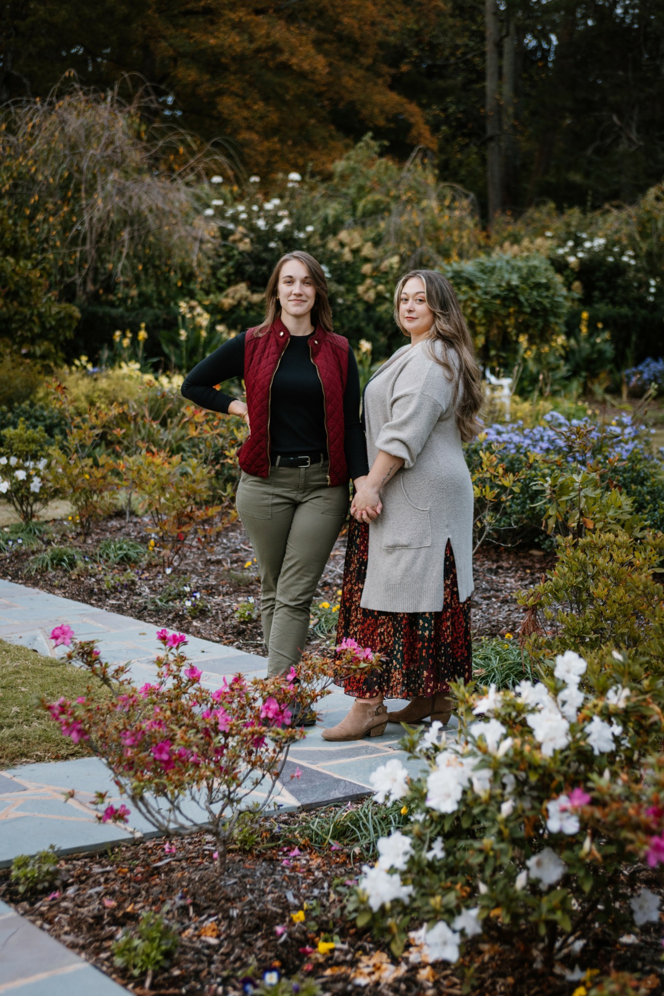 Two women stand together in a garden with colorful flowers and greenery, holding hands and smiling.