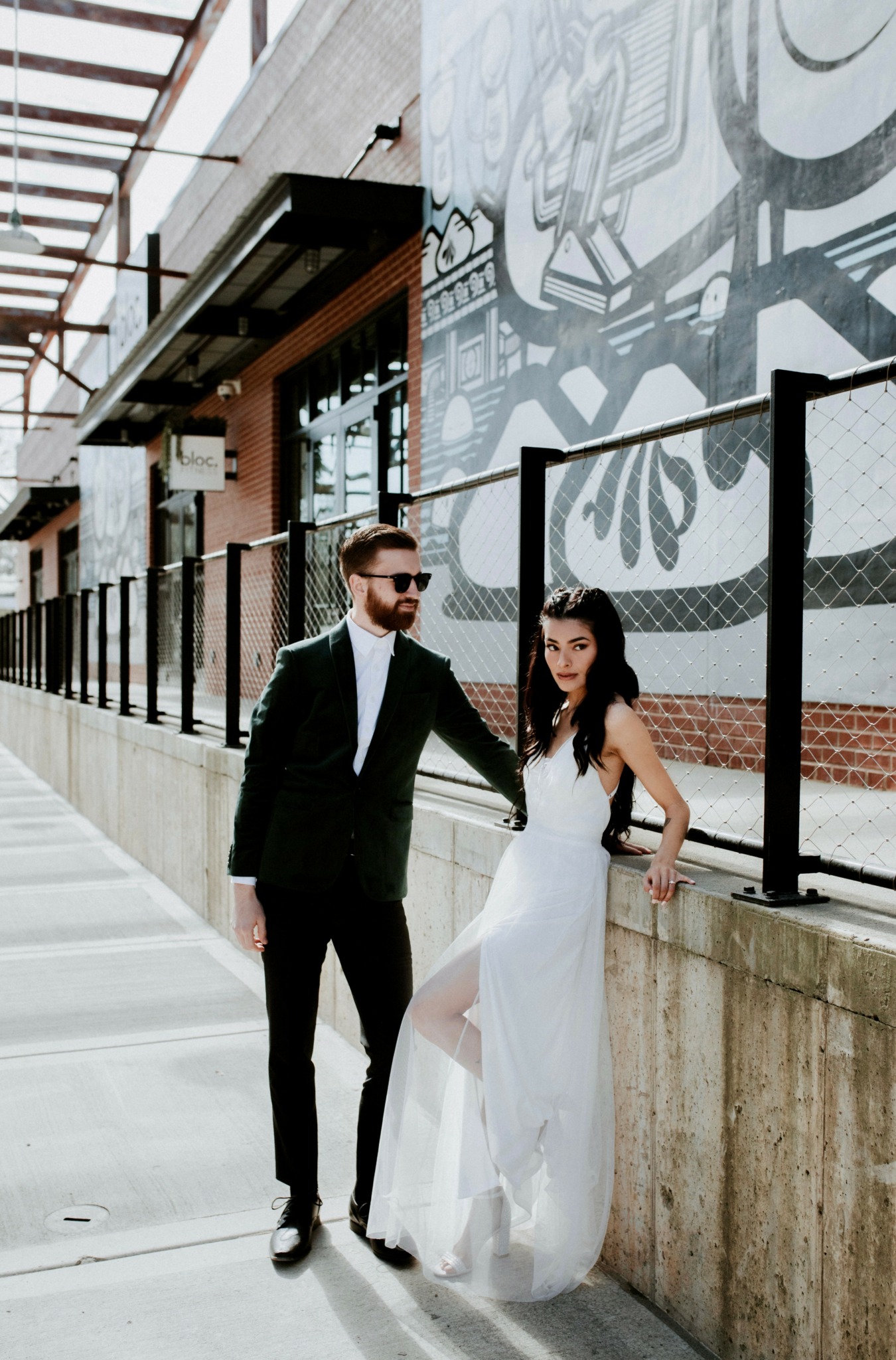Man and woman in formal attire walking along a city sidewalk with mural art on the wall behind them.