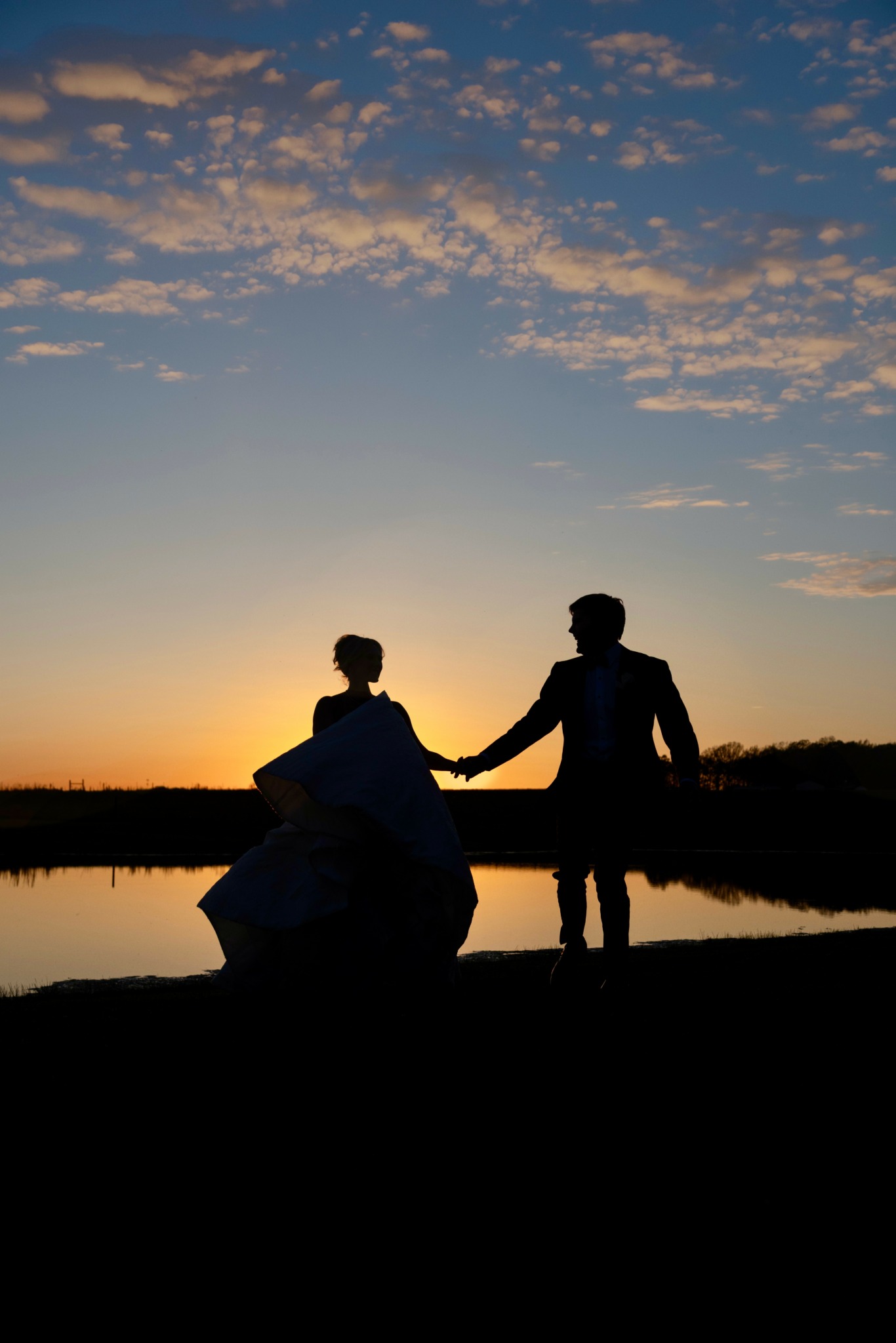 Silhouettes of two people holding hands near a body of water during sunset with a sky filled with clouds.