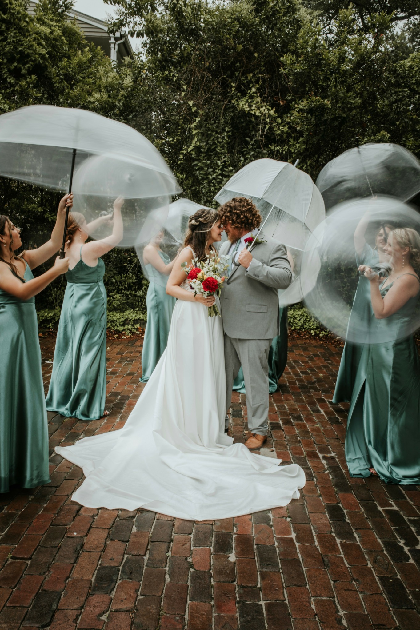 Bride and groom standing on brick path surrounded by women holding umbrellas outdoors.