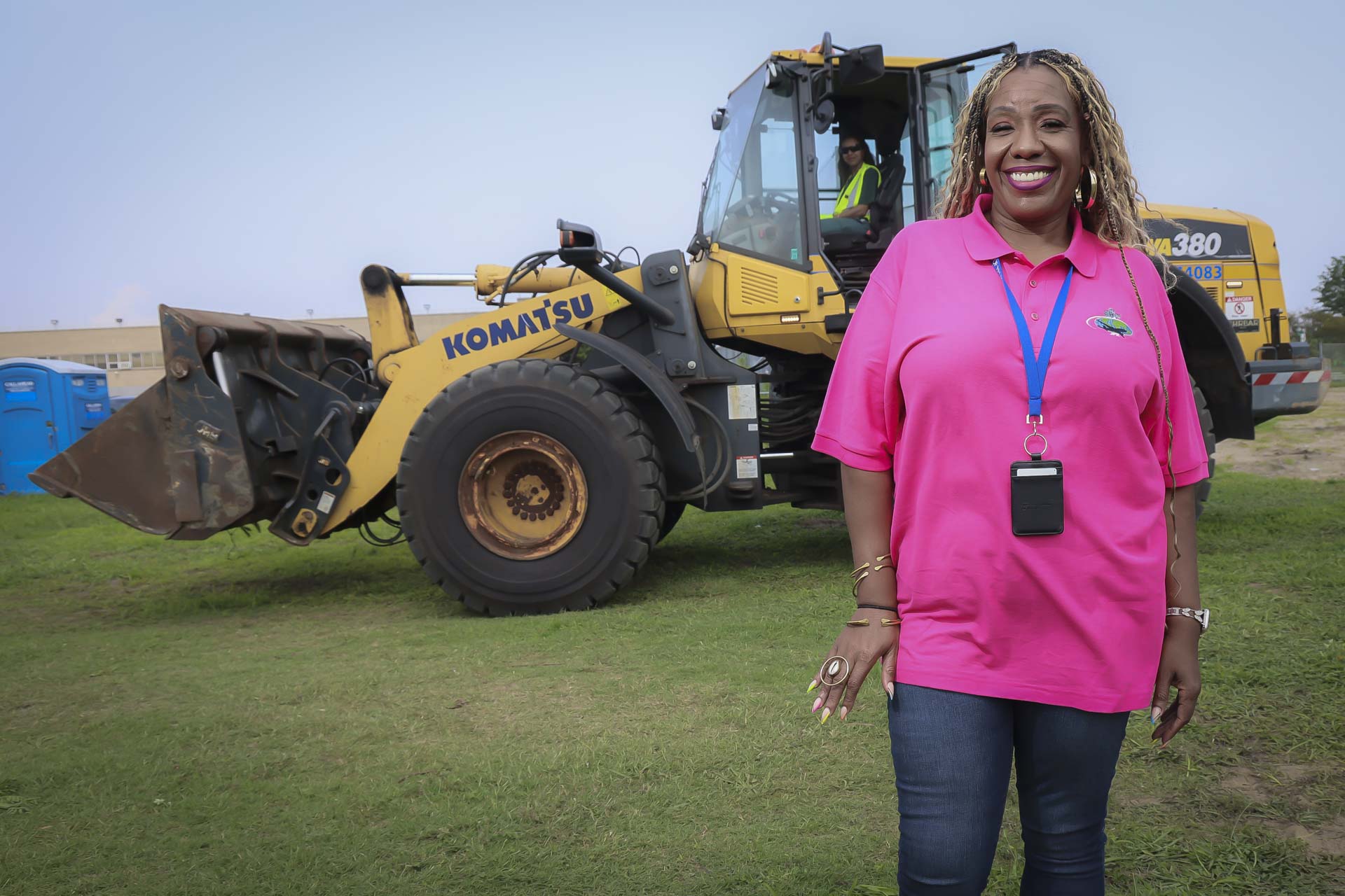 Woman in pink shirt smiling outdoors with construction vehicle in background.