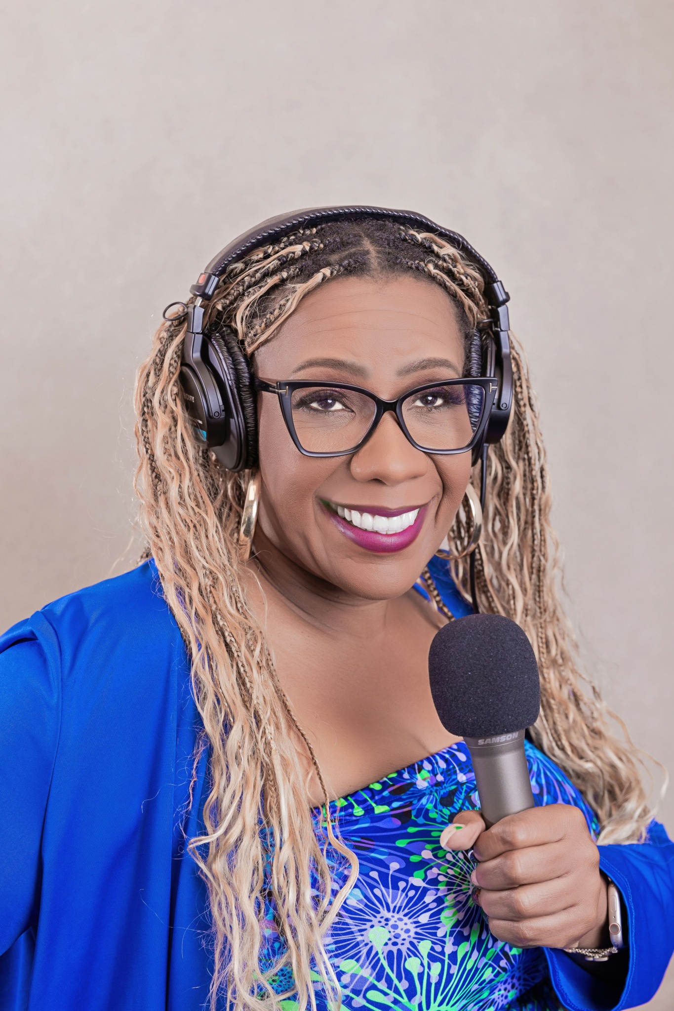 Woman with glasses, headphones, and long blonde hair holding a microphone, smiling, wearing a blue patterned top and jacket.