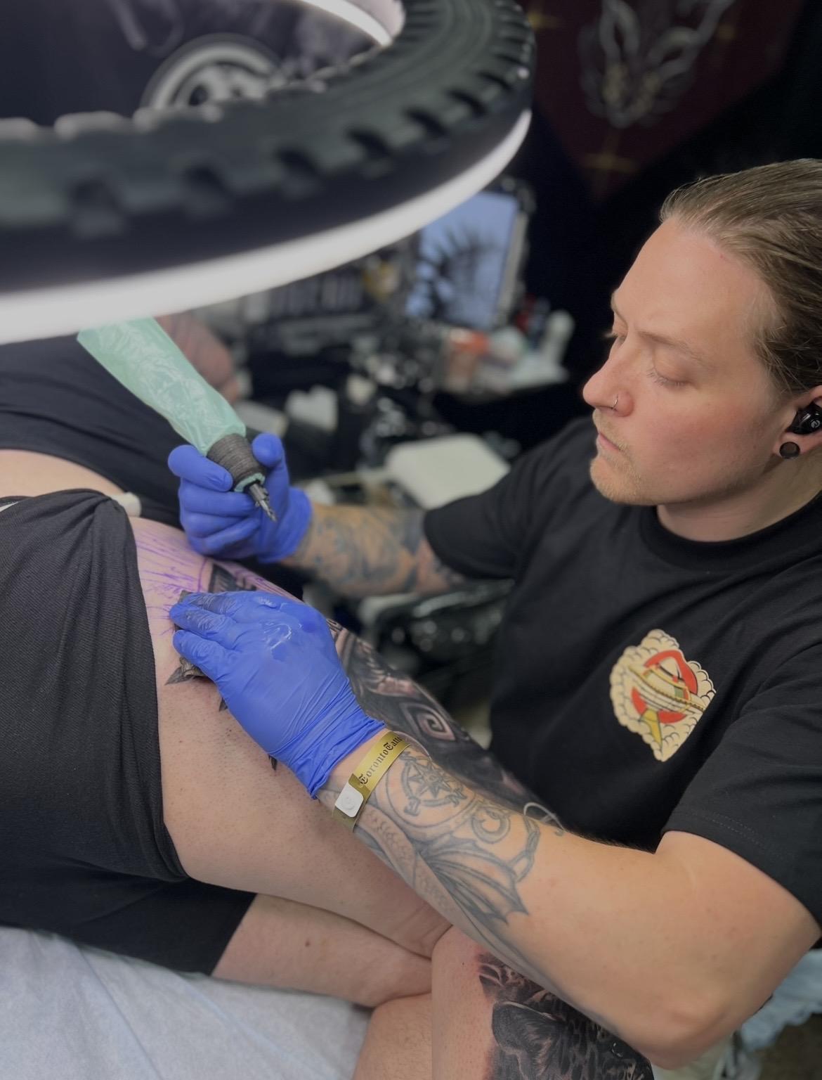 Tattoo artist wearing blue gloves working on a person's arm in a tattoo studio.